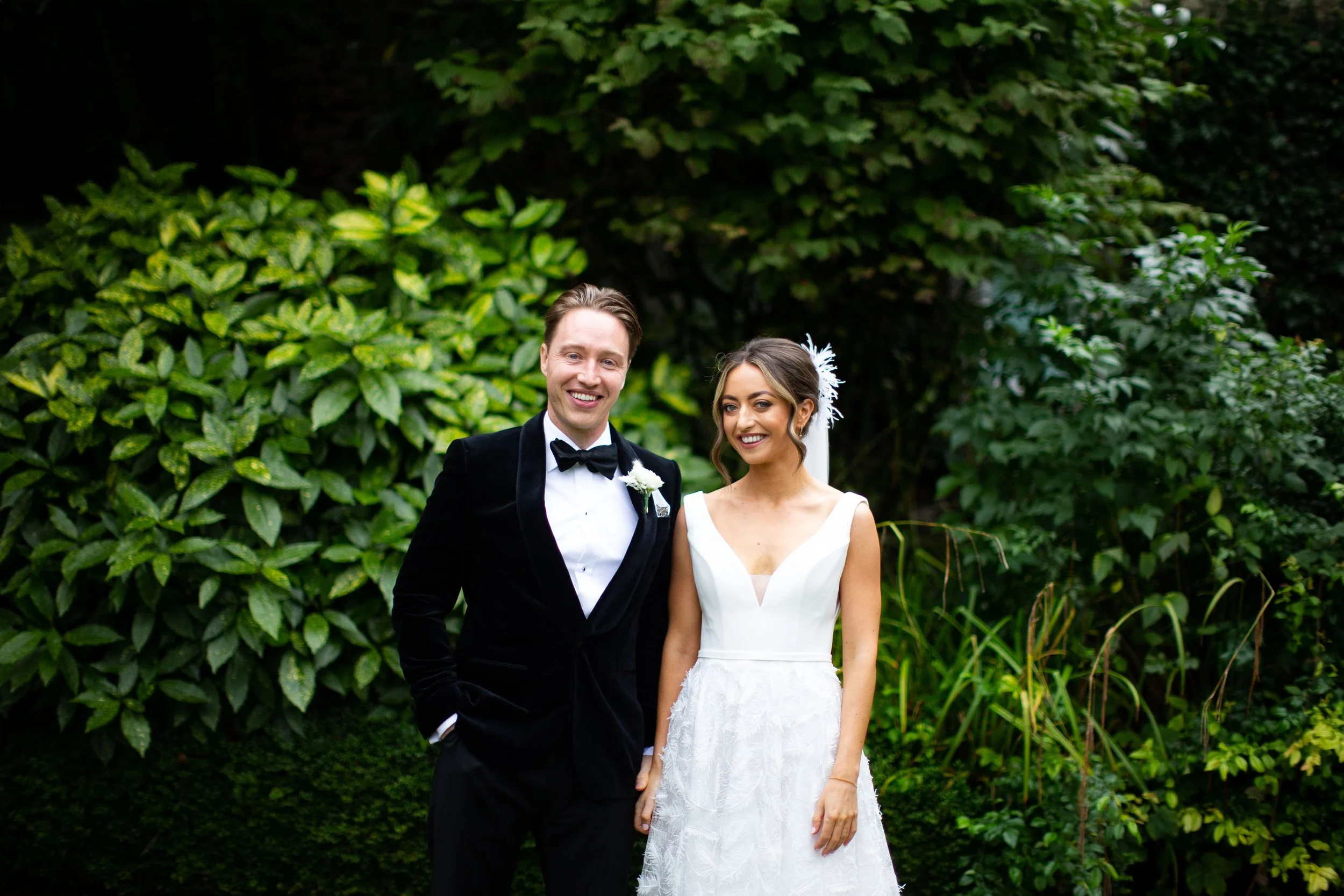 A smiling man in a black tuxedo and a woman in a white wedding dress standing outdoors in front of green bushes.