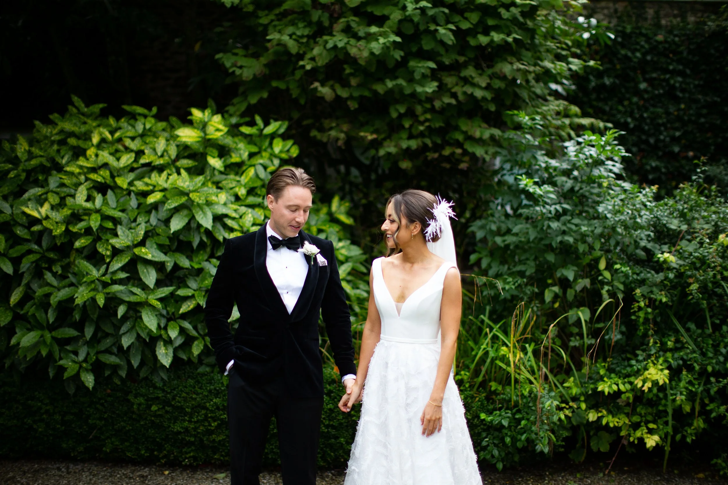 A bride and groom holding hands outdoors surrounded by greenery, dressed in wedding attire, smiling and looking at each other.