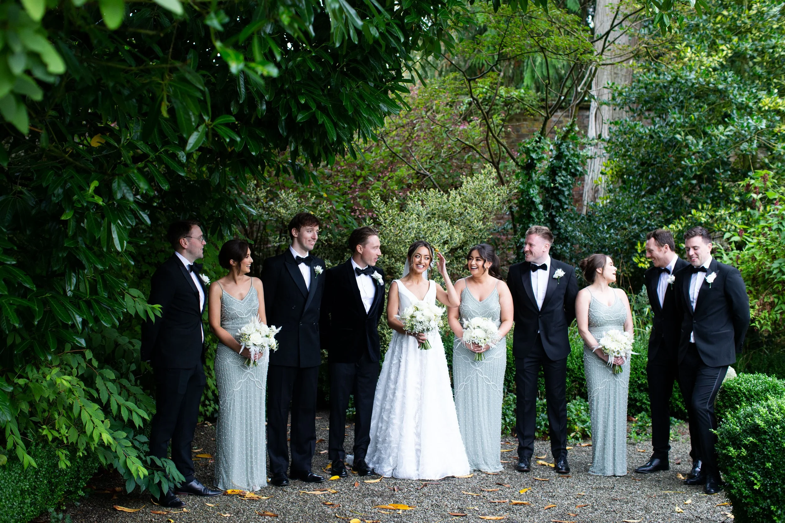 A group of wedding guests dressed in formal attire, standing outdoors in a lush green garden. The bride and groom are in the center, smiling and holding bouquets of white flowers. The guests are engaged and happy, with trees and shrubbery in the back