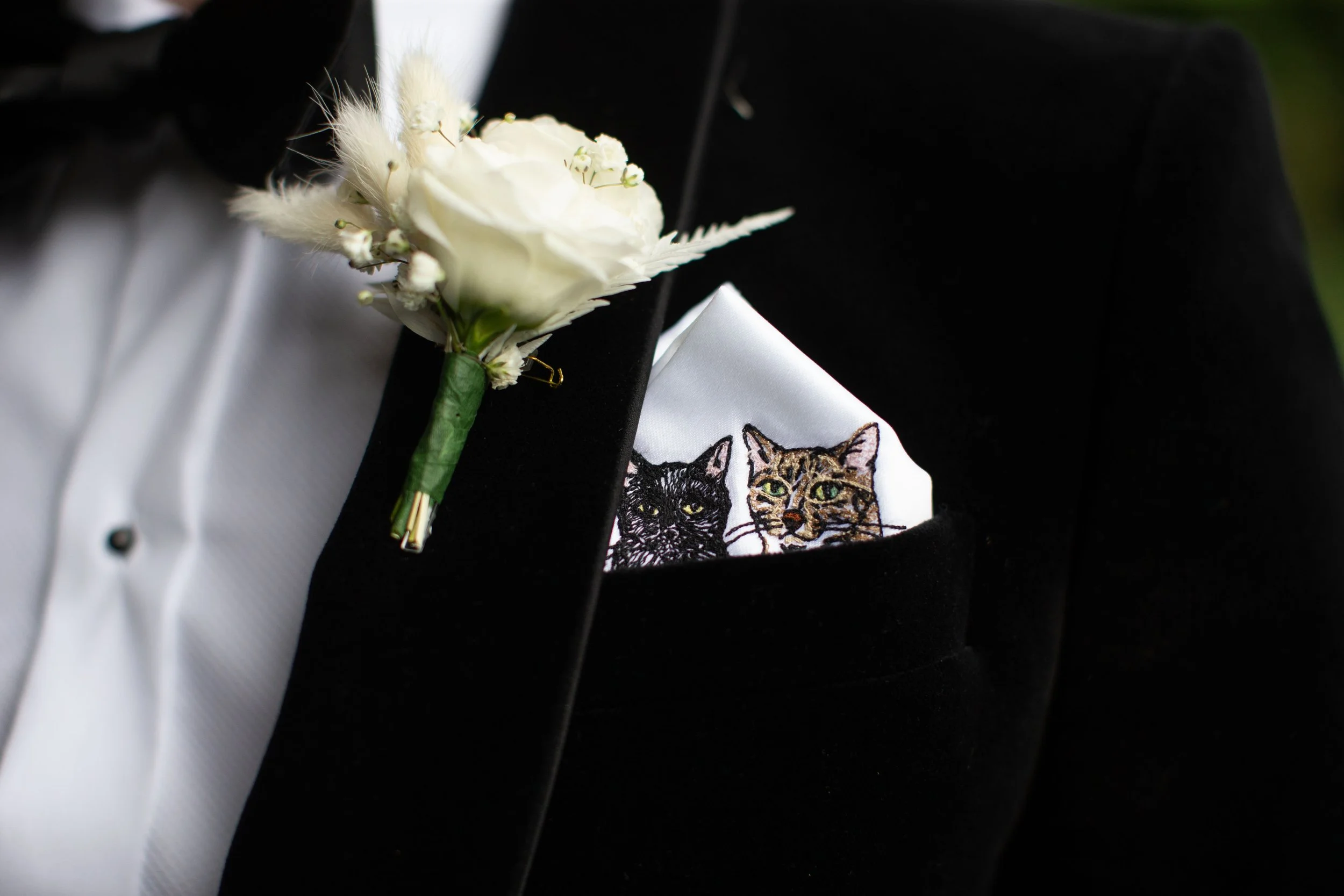 A close-up of a tuxedo jacket with a white shirt and a boutonnière made of white flowers and feathers pinned to the lapel. There is also a pocket square with cat illustrations peeking out of the pocket.