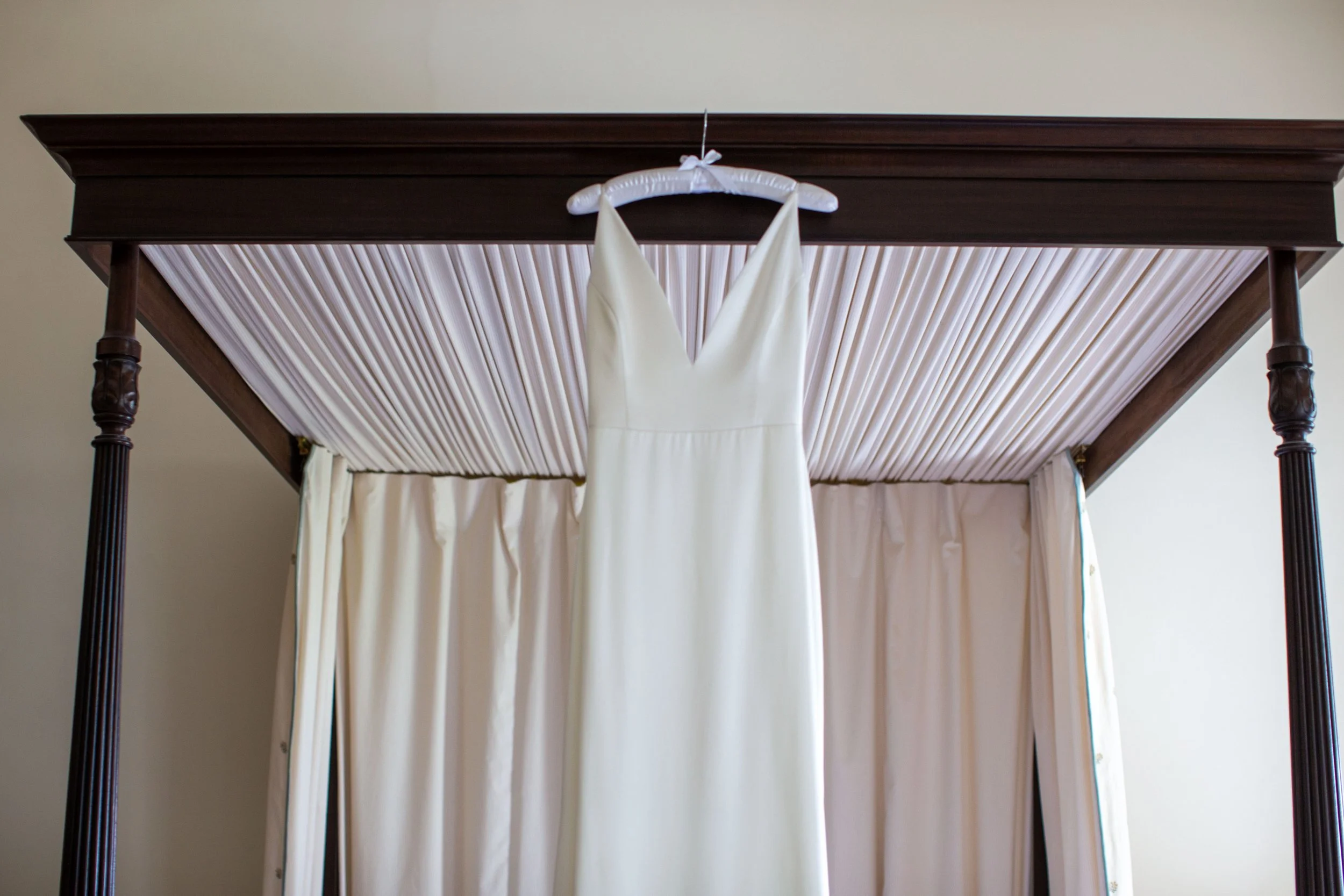 A white wedding dress hanging on a padded hanger from a four-poster bed with dark wooden posts and a canopy, inside a room with cream-colored walls.