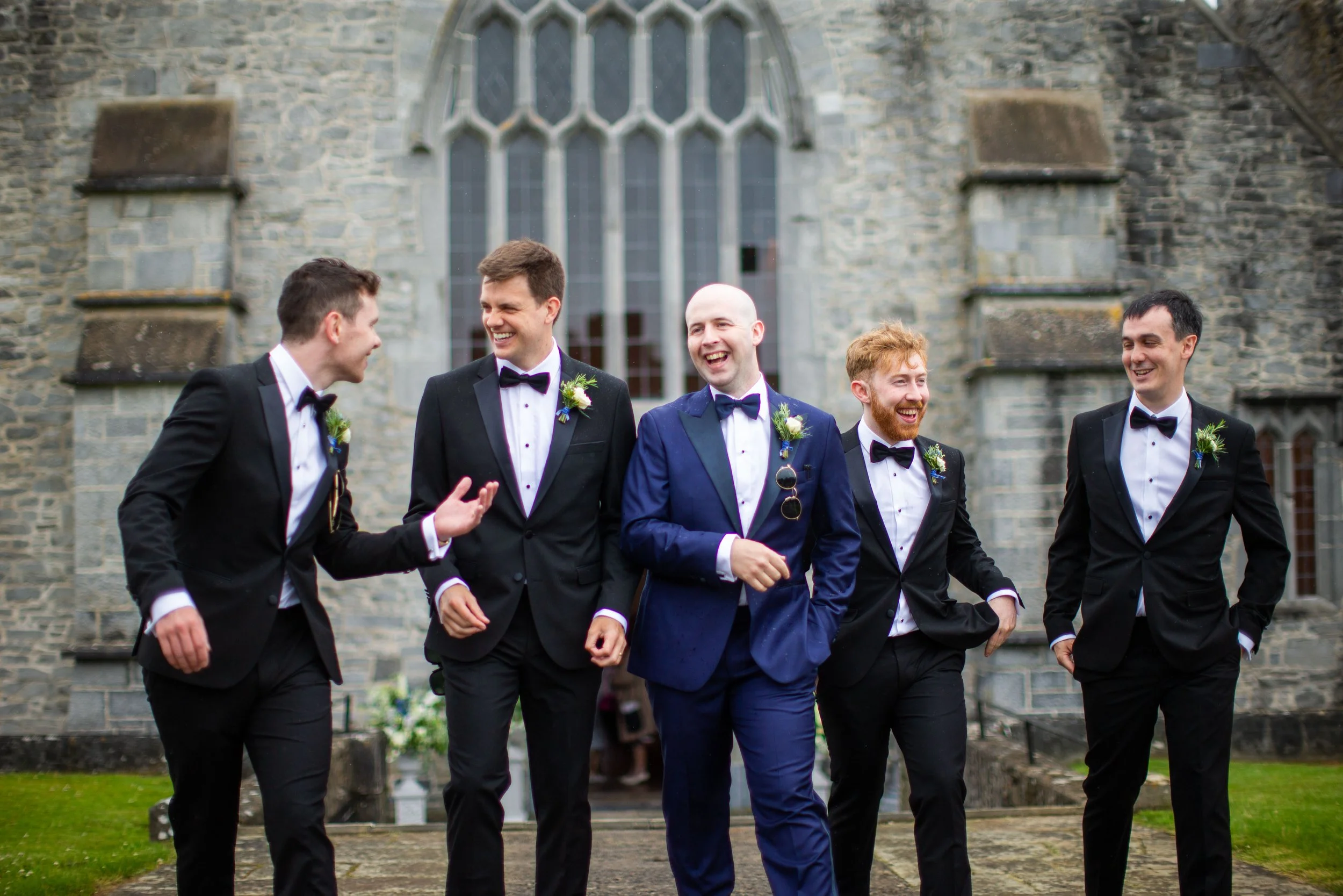 A group of five men in tuxedos and bow ties, talking and smiling, walking outside a stone church with large windows, during a wedding celebration.
