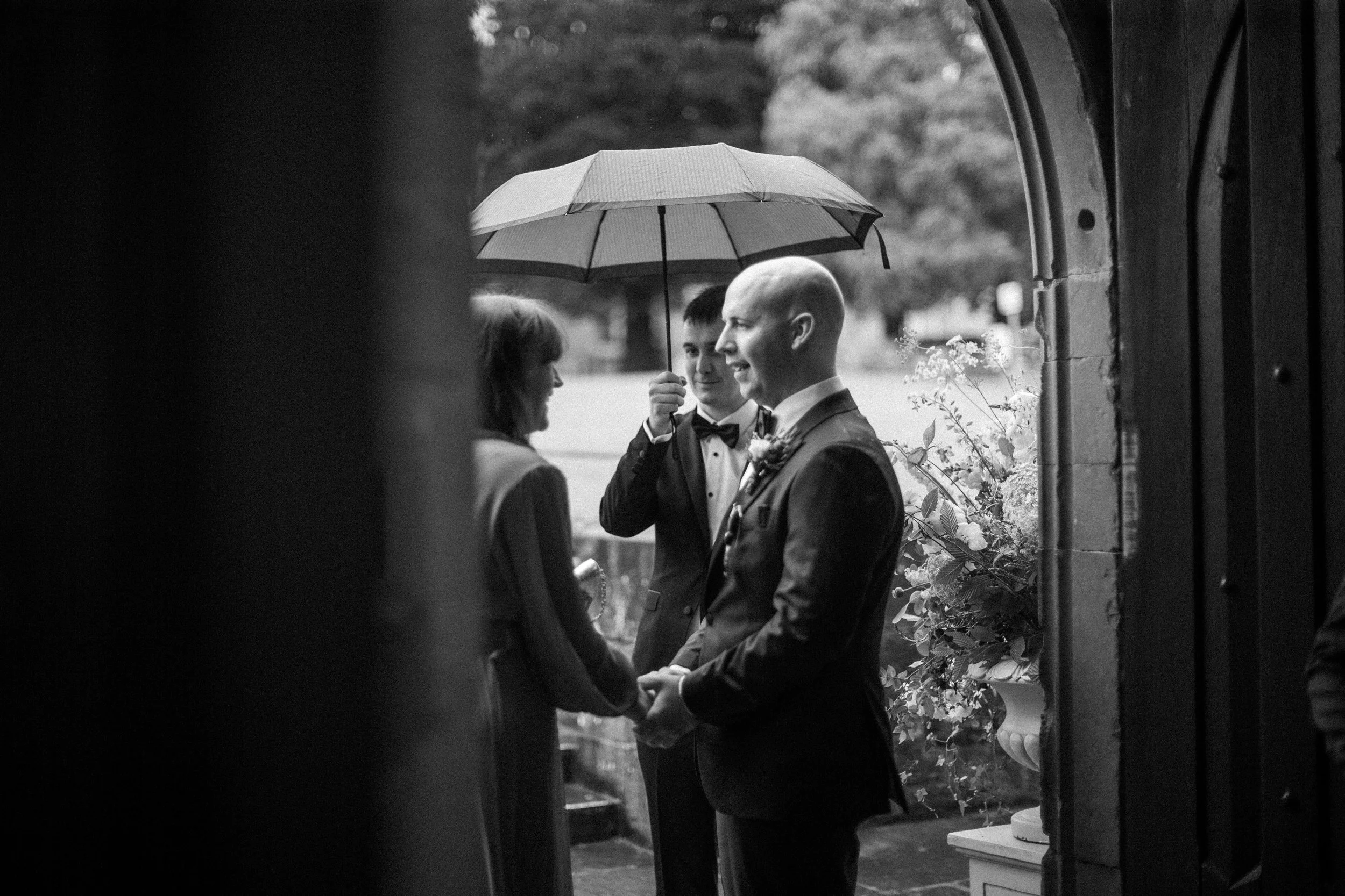 A black-and-white photograph of a wedding ceremony, seen from inside a building through an archway. A bride and groom hold hands and gaze at each other, with a person holding an umbrella over them. The background features trees and a garden.