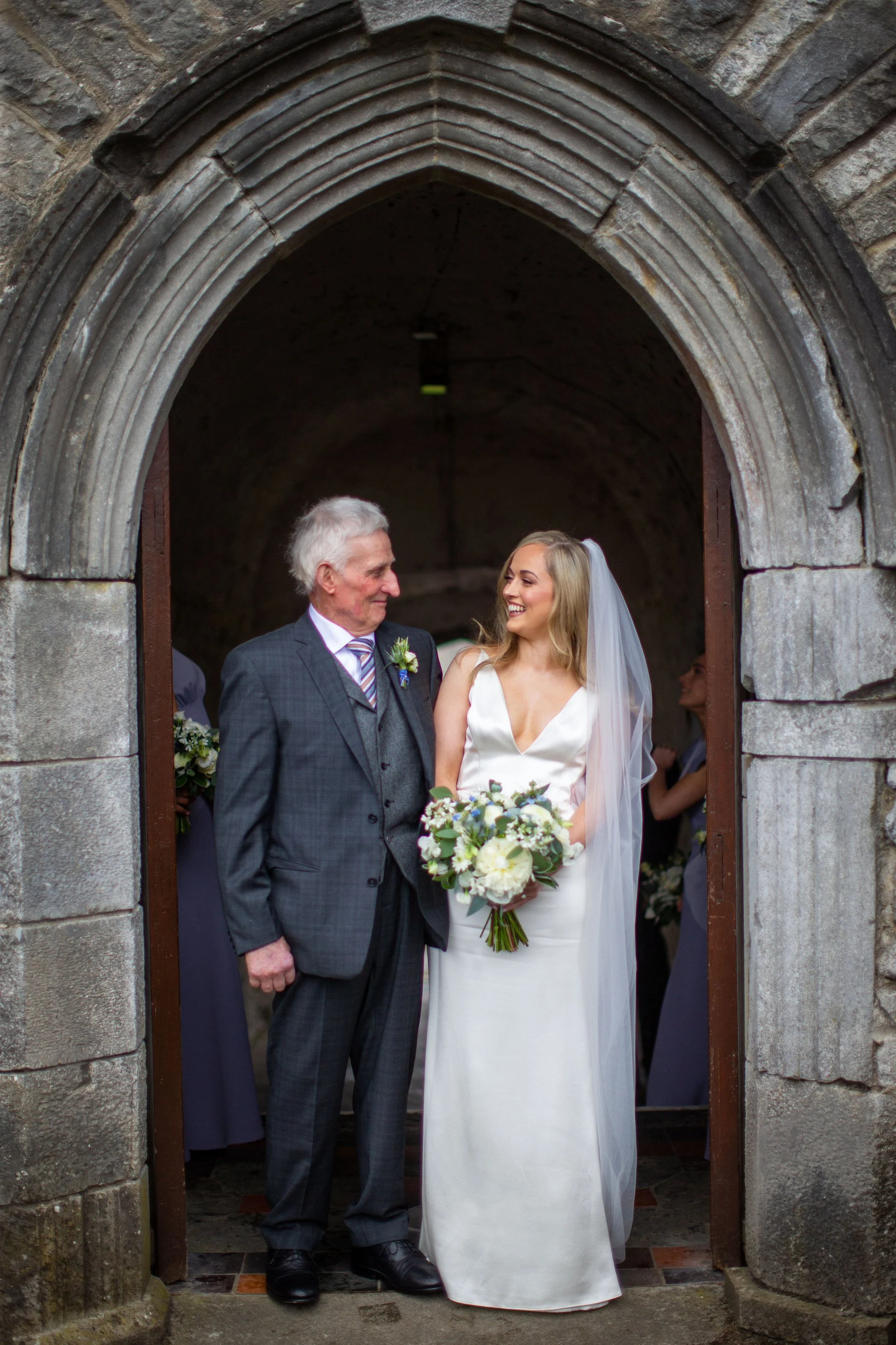 A bride in a white wedding dress holding a bouquet, smiling at an older man in a gray suit and tie, standing within a stone archway of a church.