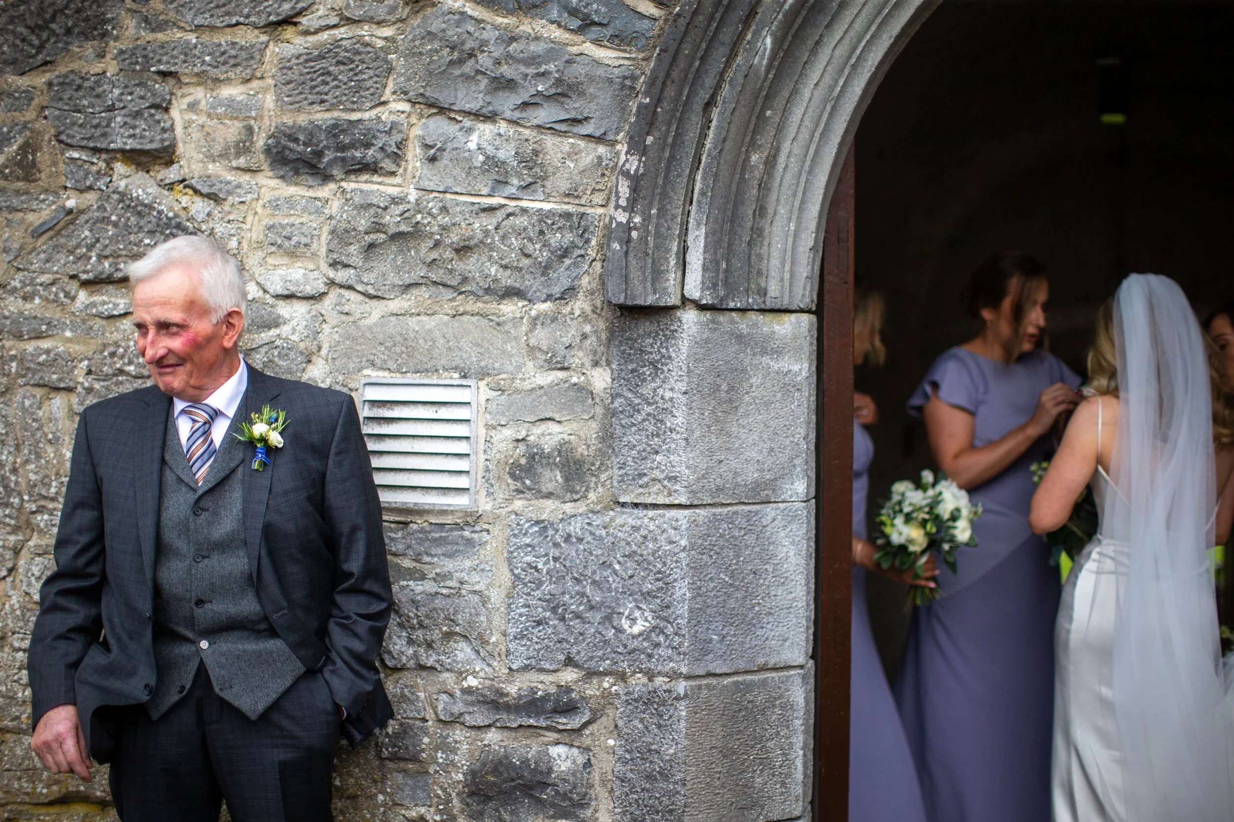An elderly man in a suit with a boutonniere, standing outdoors against a stone wall, while a group of women, including a bride with a veil and bouquet, are visible through an arched doorway in the background, likely at a wedding.