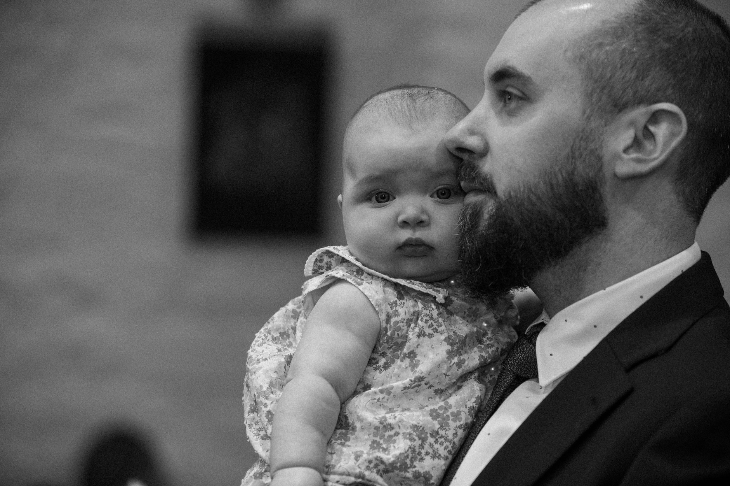 A man with a beard holding a young girl with curly hair, both dressed in formal attire. The girl looks directly at the camera, while the man's face is in profile, looking away.
