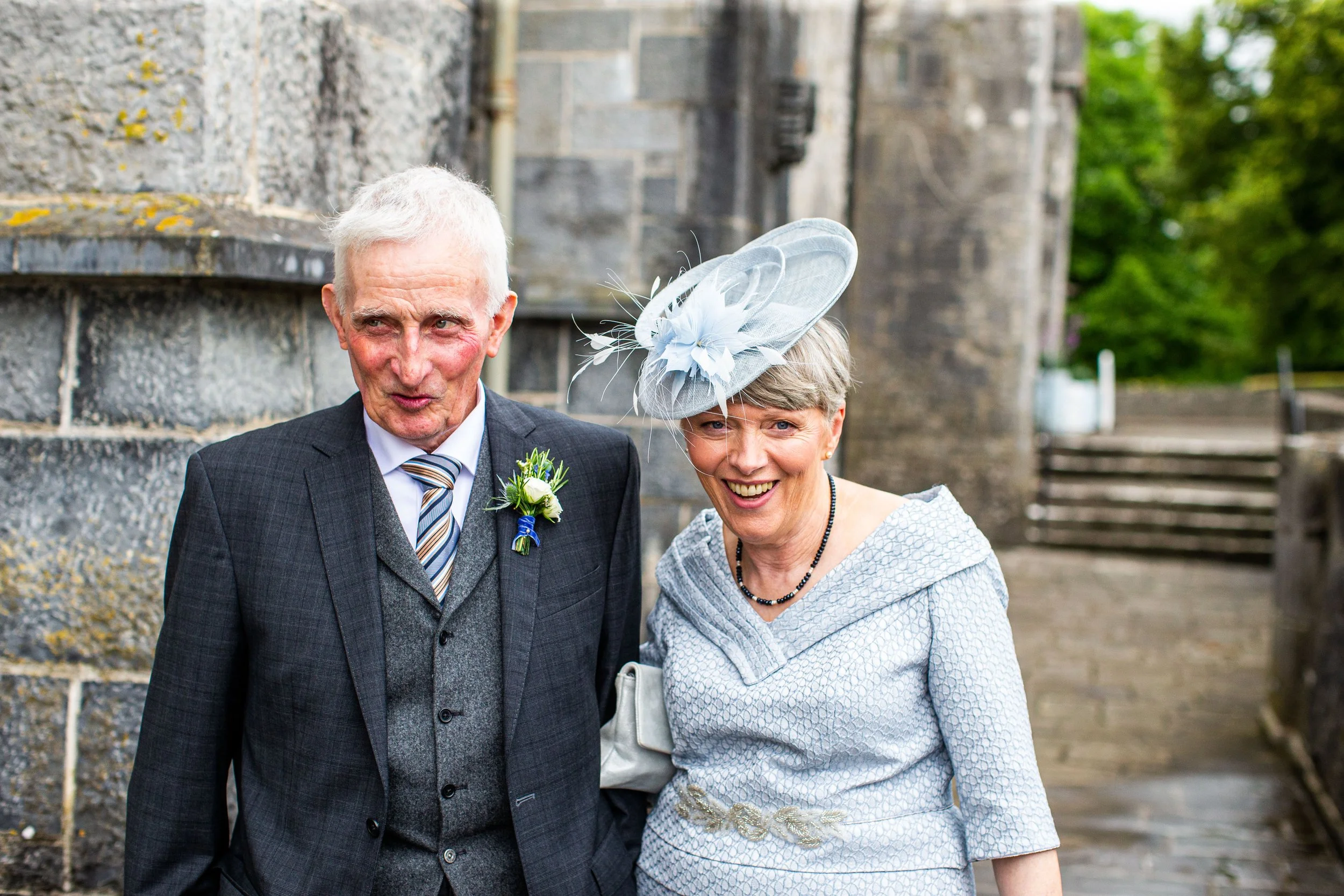 An elderly man in a suit and a woman wearing a fancy hat stand together outdoors near a stone wall.