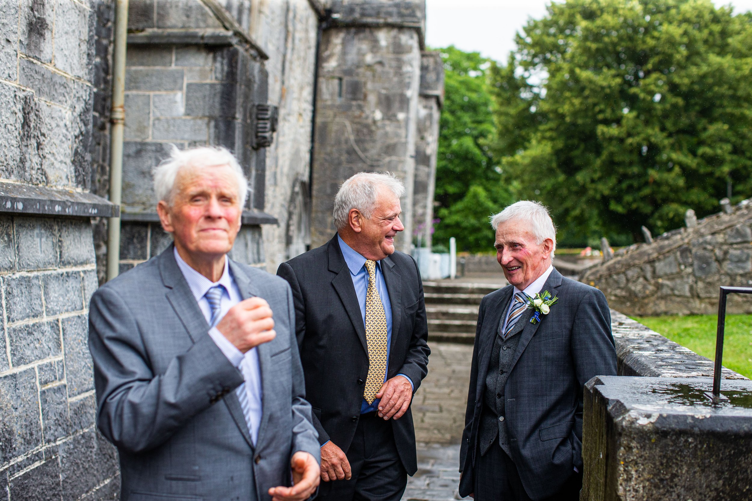 Three older men dressed in suits standing outside near a stone wall, engaged in conversation and smiling, with green trees in the background.