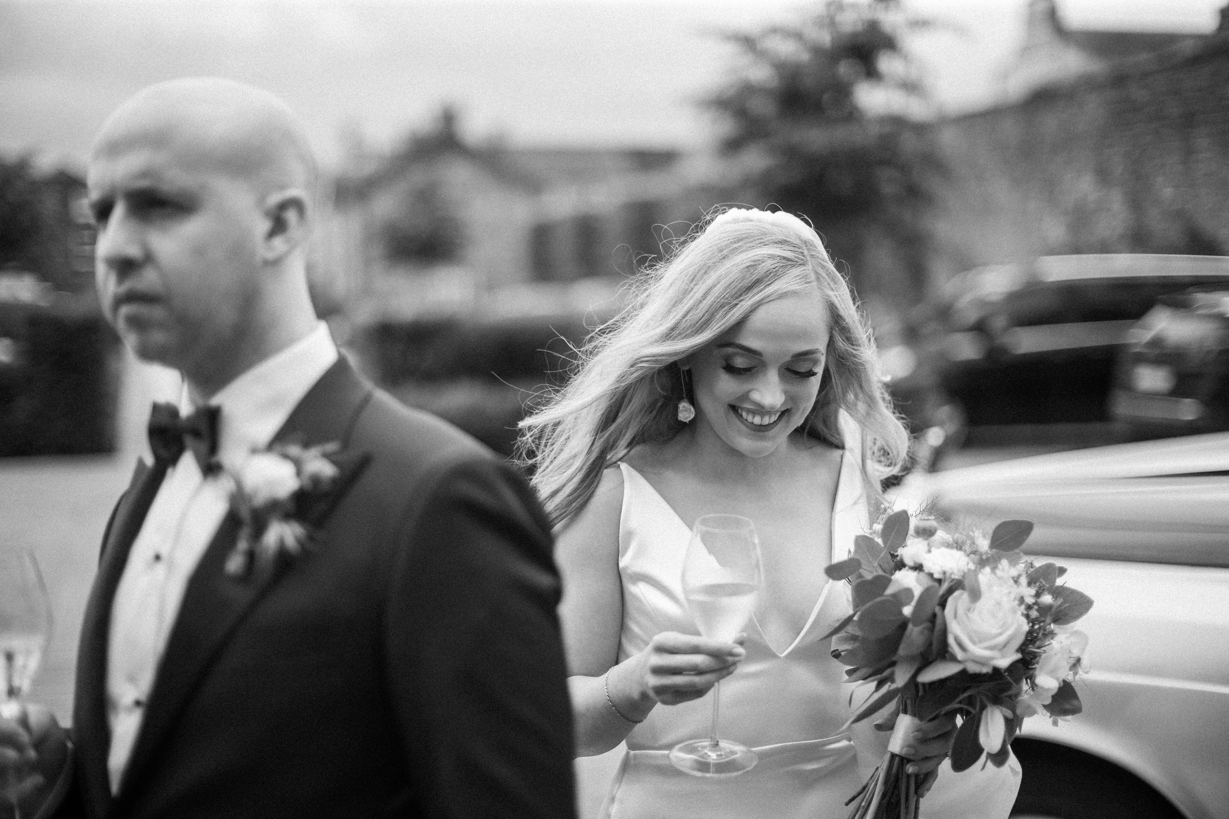 Black and white photo of a woman in a wedding dress holding a bouquet of flowers, smiling and reading a card, while a man in a tuxedo stands nearby. The background appears to be outdoors with cars and trees.