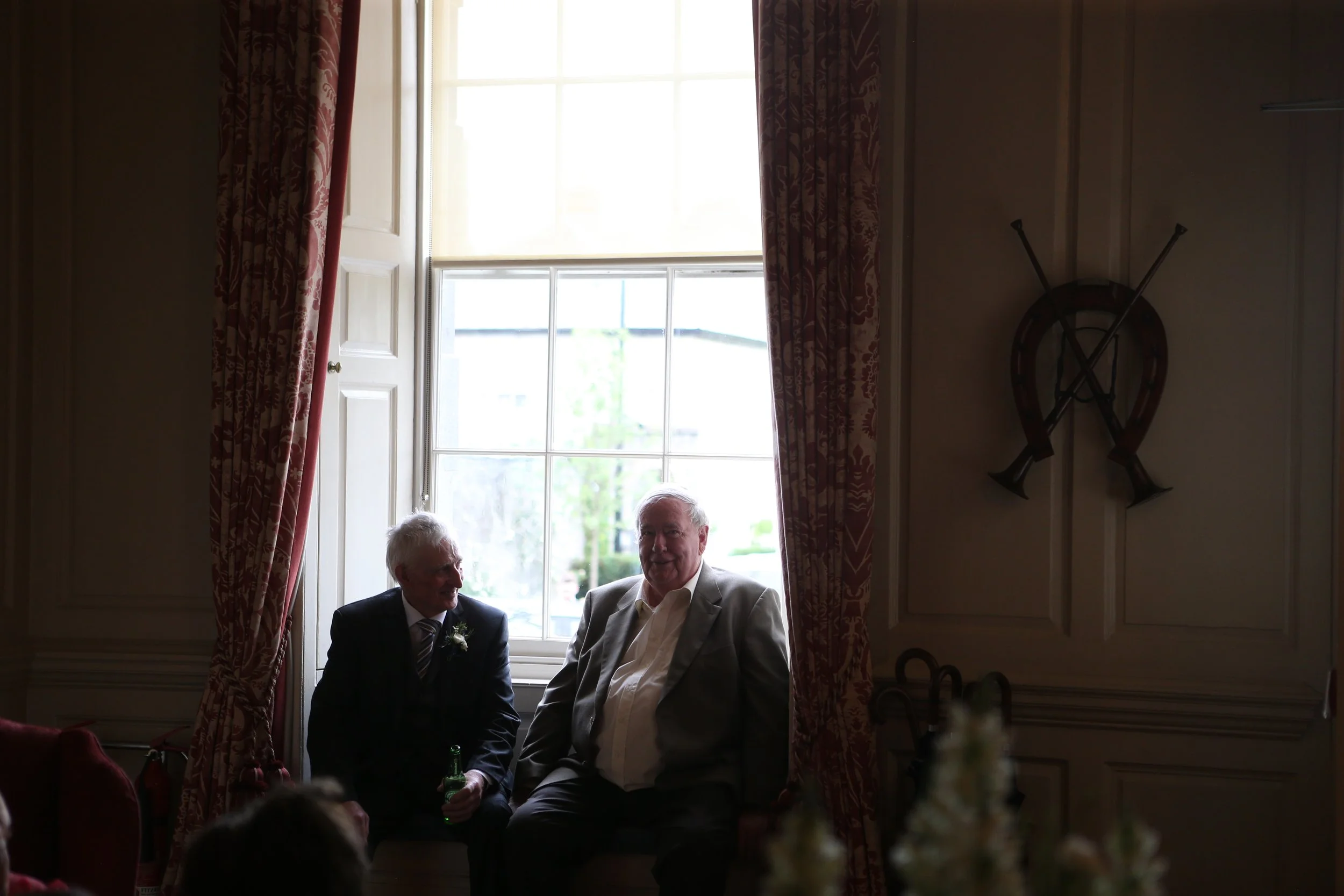 Two men sitting in a room near a large window with floral curtains, engaged in conversation. One man has white hair and is holding a green bottle, while the other has gray hair and is smiling. Sunlight streams in through the window, brightening the s