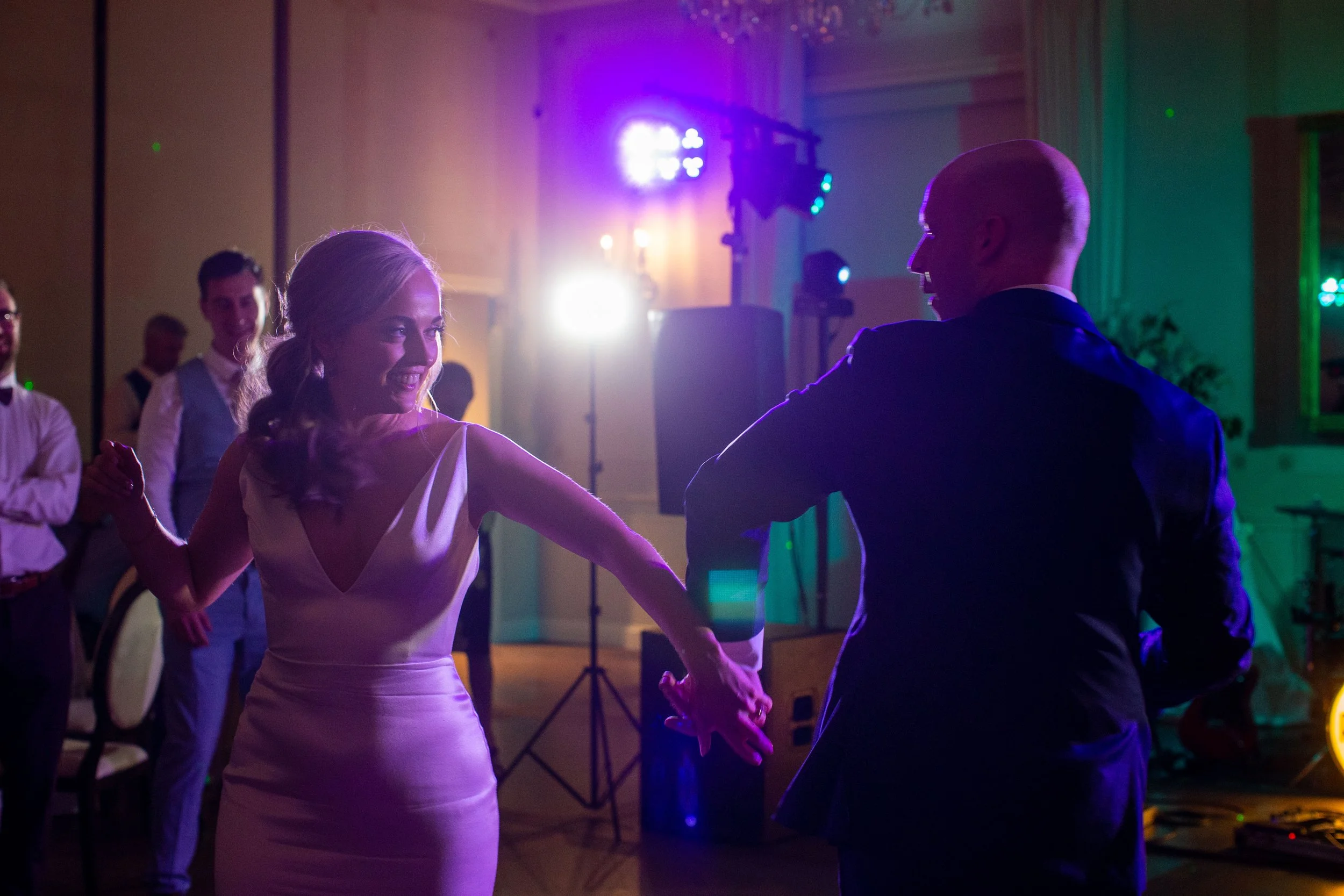 A woman in a white dress dancing with a man in a suit at a social event, with other guests and colorful lighting in the background.