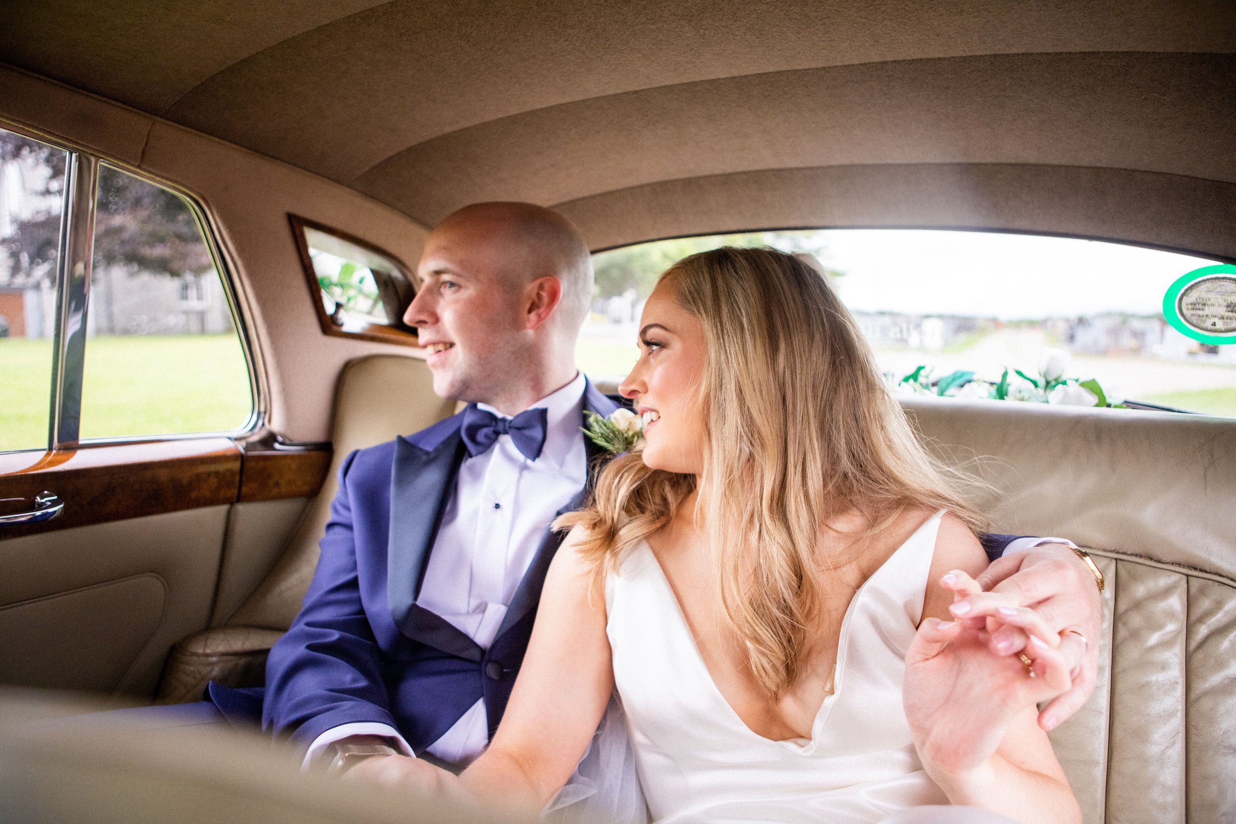 A bride and groom sitting together in the backseat of a vintage car during their wedding, holding hands and smiling.
