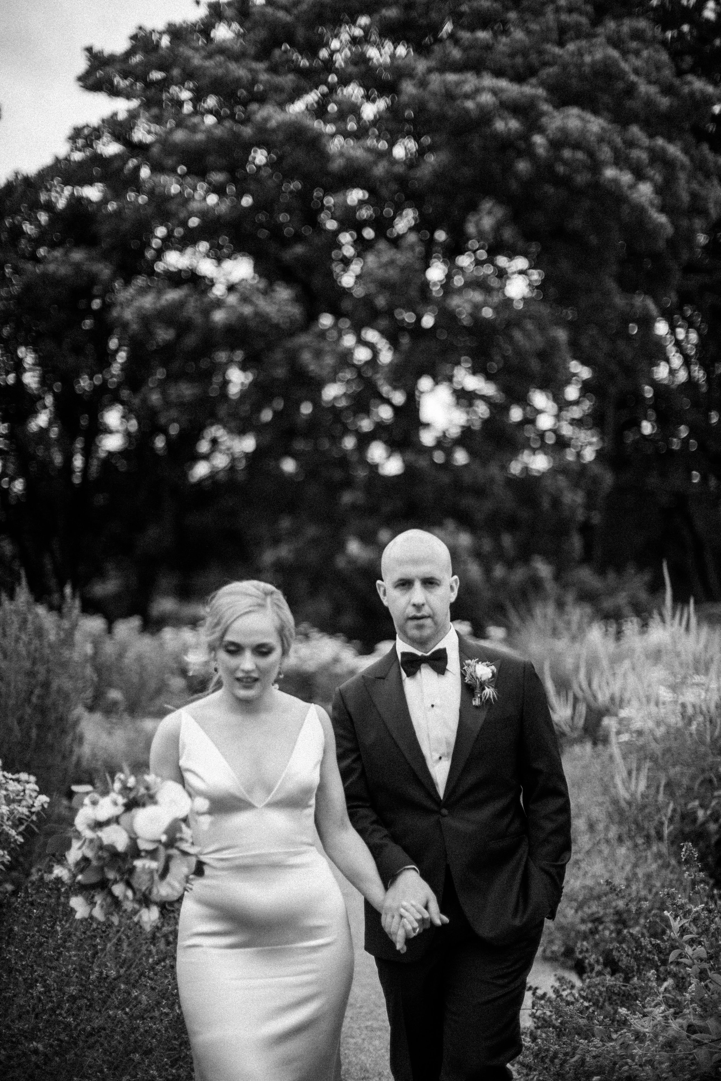 A black and white photo of a bride and groom walking outdoors hand in hand, surrounded by tall grass and trees, during a wedding. The bride is wearing a white wedding dress and holding a bouquet of flowers. The groom is dressed in a black tuxedo with