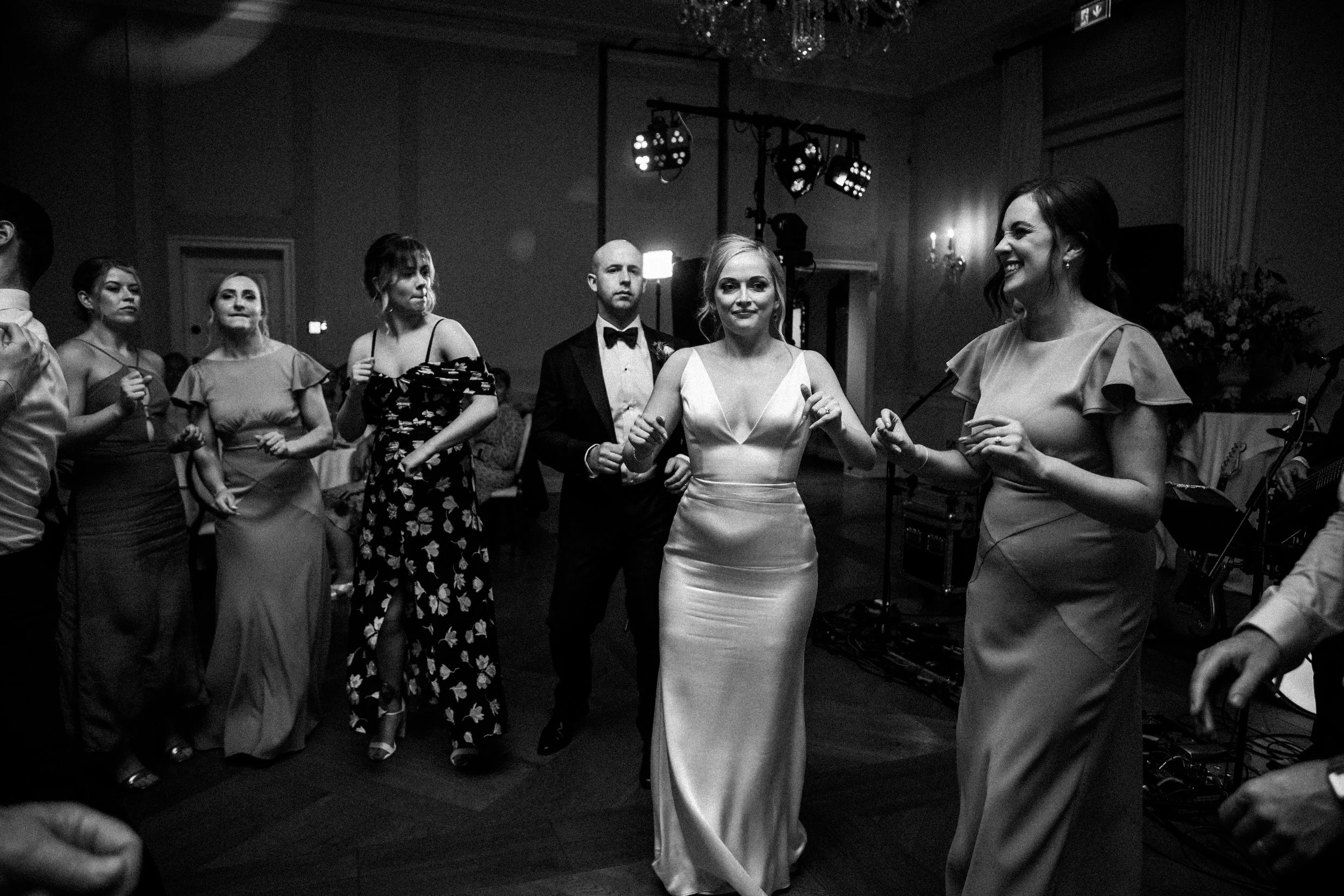 A black and white photo of a group of people at a wedding reception. The bride is in the center, wearing a satin wedding gown, dancing with a woman in a ruffle dress on her right. A groom in a tuxedo with a bow tie stands behind the bride. Other wome