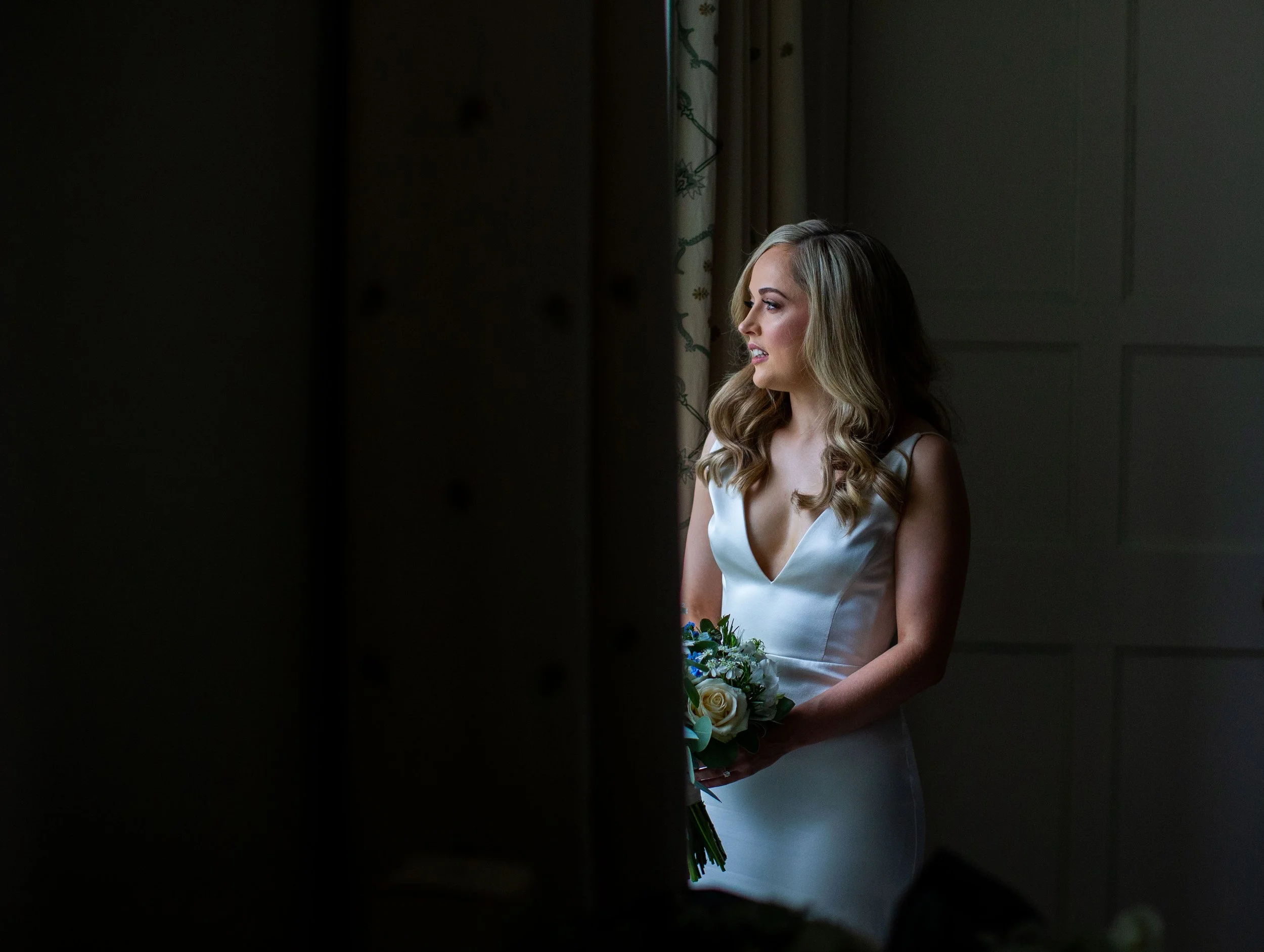 A woman in a white dress holding a bouquet of white roses and greenery, standing in front of a window with floral curtains, looking to her left in a softly lit room.