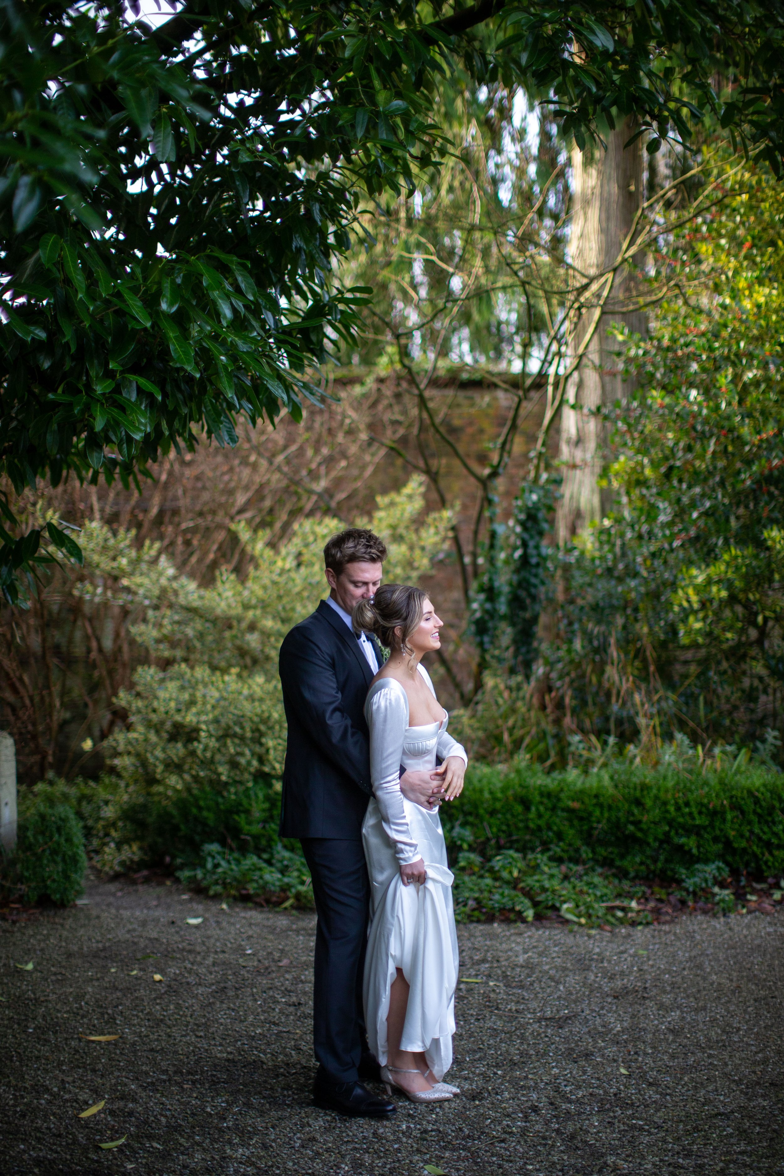 A man in a black suit and a woman in a white dress are standing outdoors in a garden, embracing each other and smiling. There are green trees and bushes in the background.