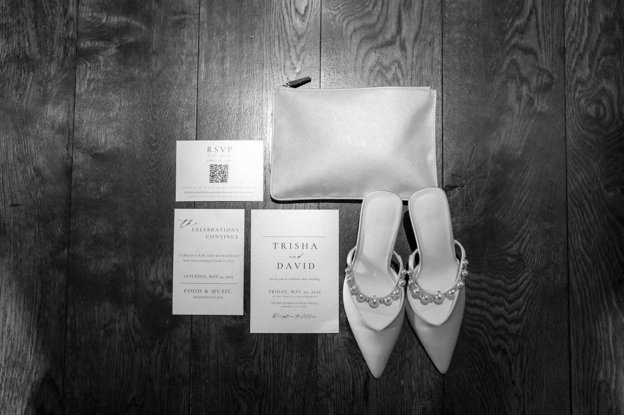 A wedding invitation suite, a pair of white bridal shoes with pearl details, and a silver clutch purse arranged on a dark wooden floor. The invitation includes details such as RSVP, date, location, and bride and groom's names, Trisha and David.