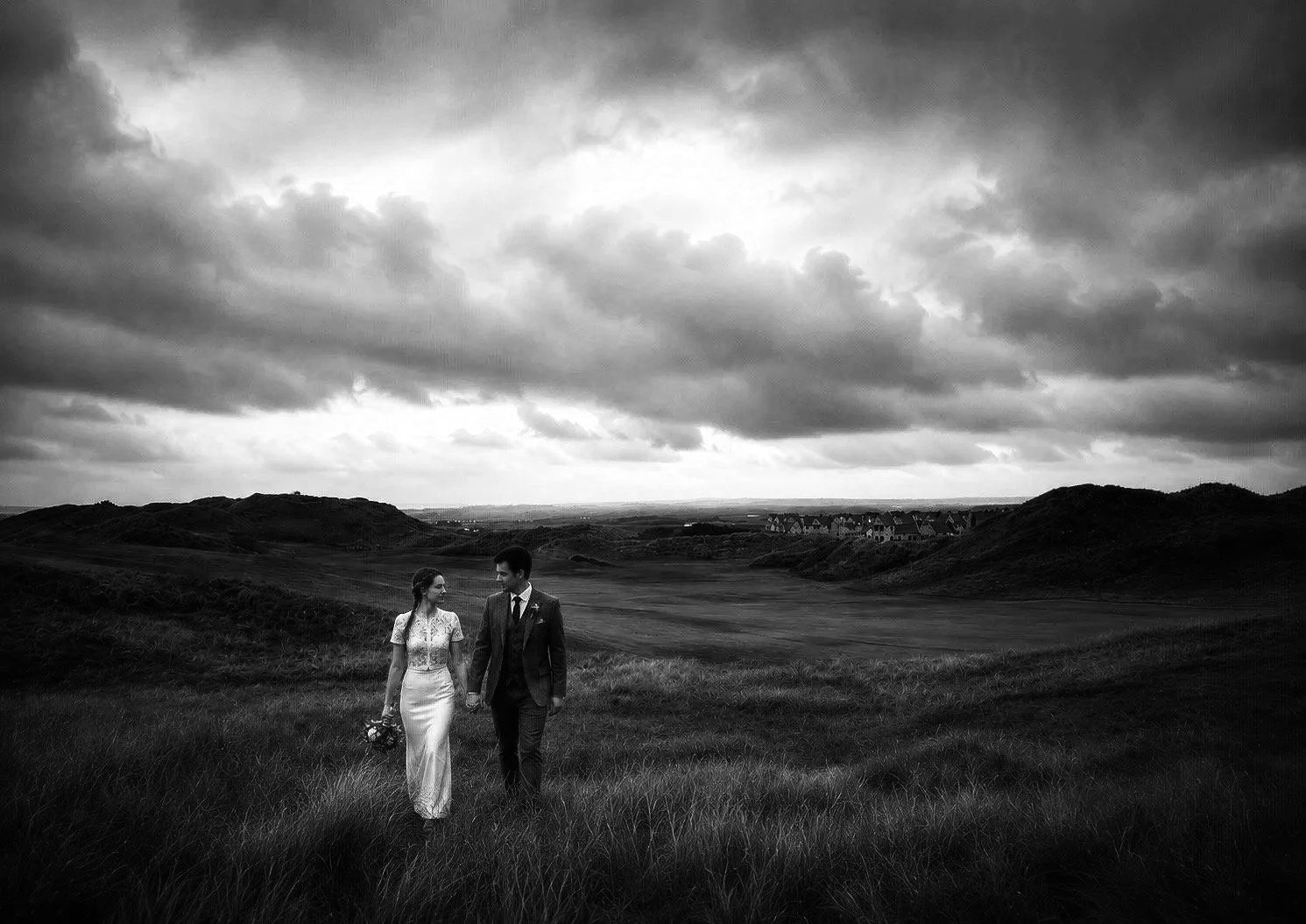 A black and white photo of a bride and groom walking hand in hand across a grassy field with rolling hills and dramatic cloudy sky in the background.