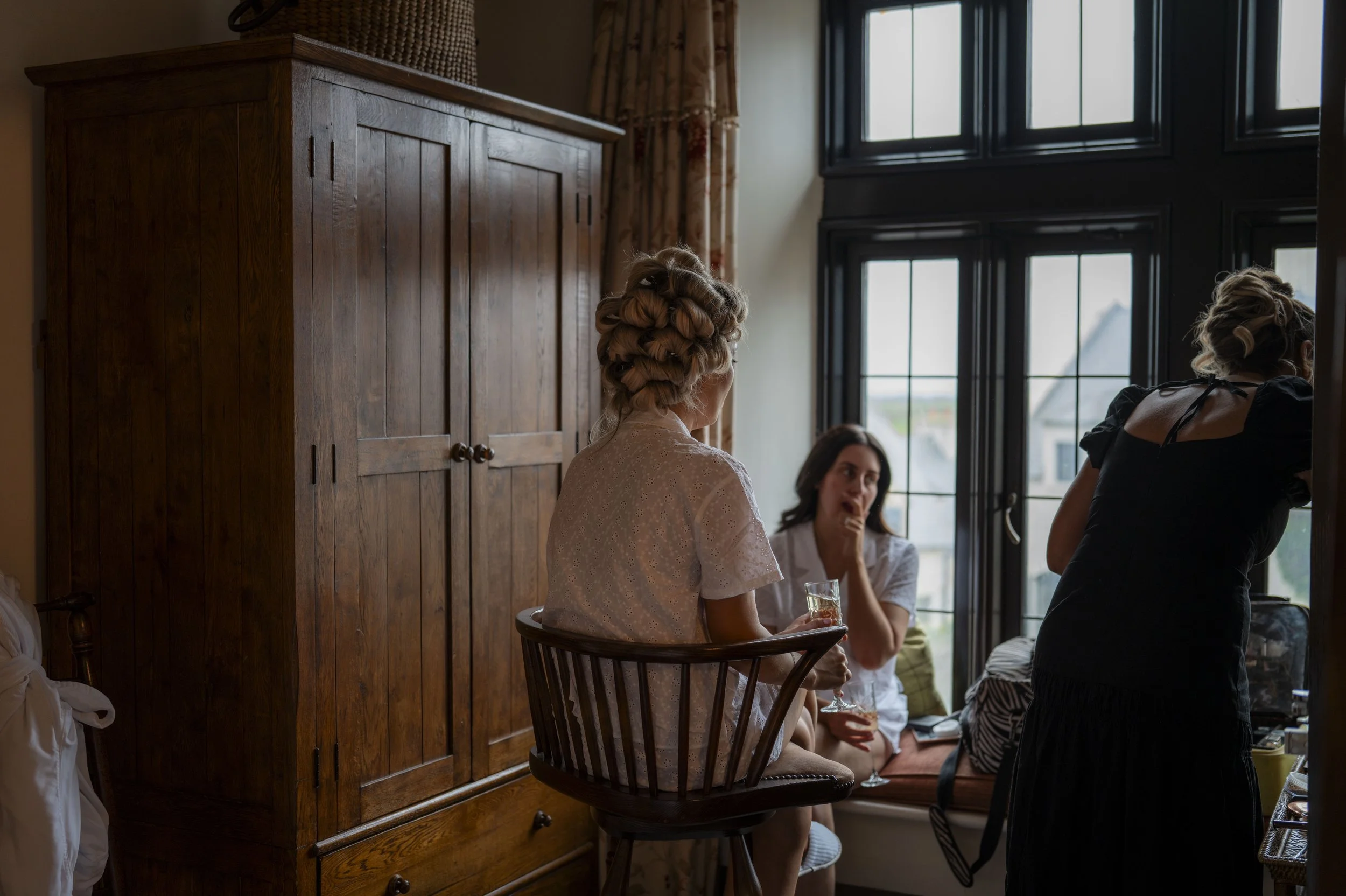 A group of women gathered in a room by a large window, with one woman seated on a wooden chair wearing a vintage-style dress with her hair styled in curls, and others standing and sitting nearby, holding drinks and engaging in conversation.