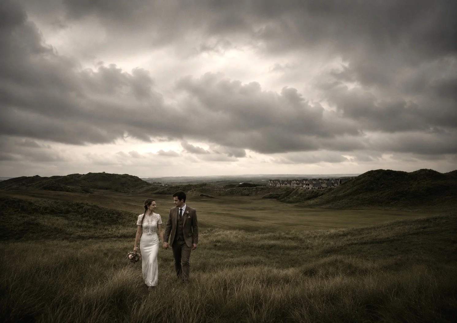 A bride and groom walking hand in hand across a grassy field under dark, cloudy skies with rolling hills in the background.