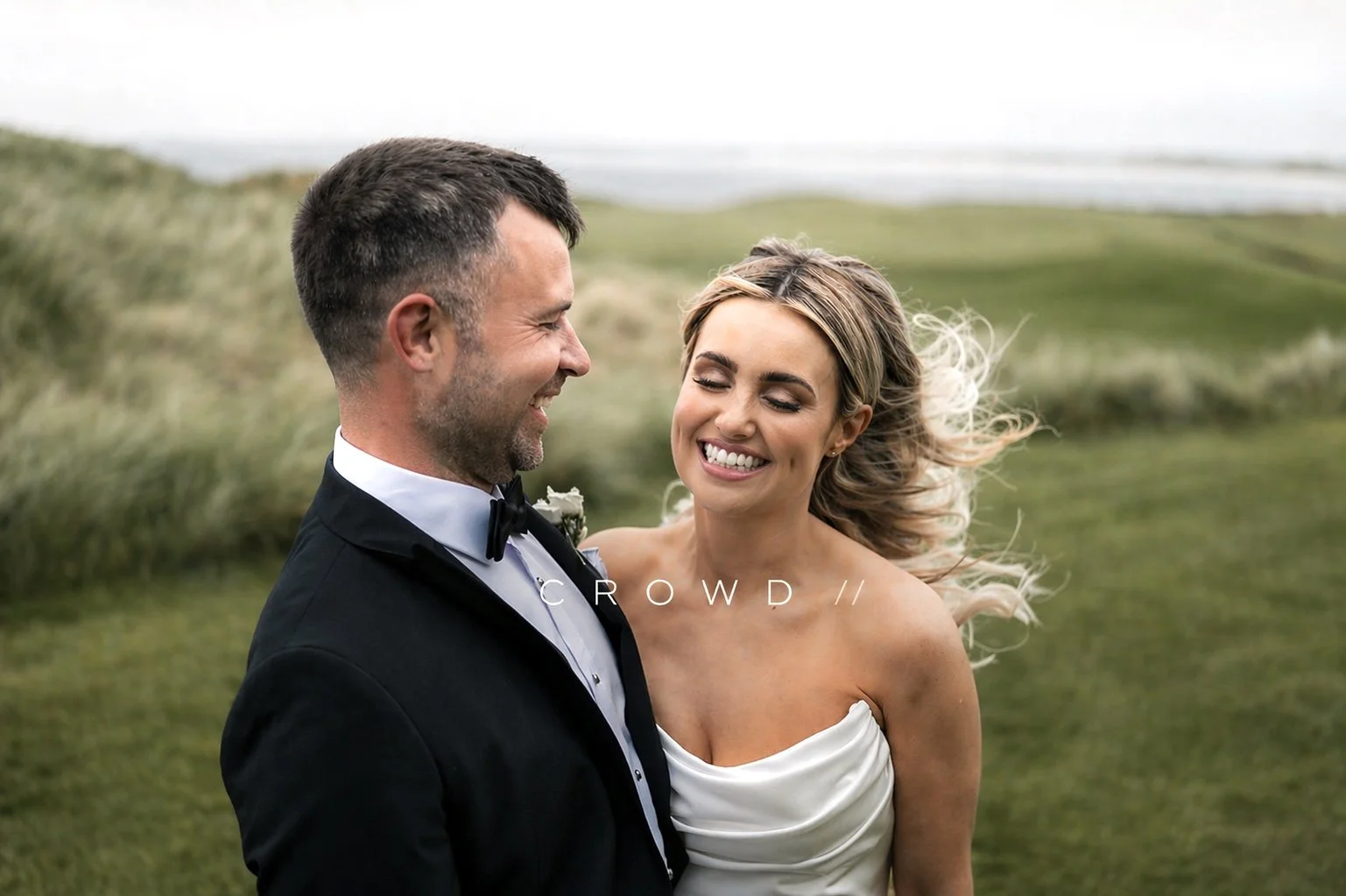 A smiling bride and groom in wedding attire standing outdoors in a green, natural landscape with grass and distant hills.