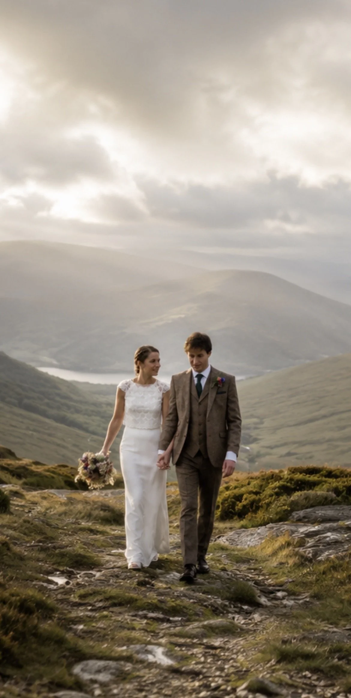 A bride and groom walking hand in hand outdoors in a scenic landscape with mountains and cloudy sky.