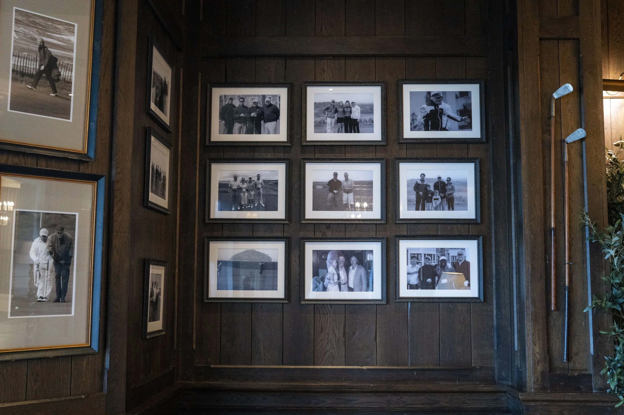 A display of framed black and white photographs on a dark wooden wall, featuring people, some dressed in golf attire, standing outdoors. The photographs are arranged in a grid pattern with some frames larger than others.