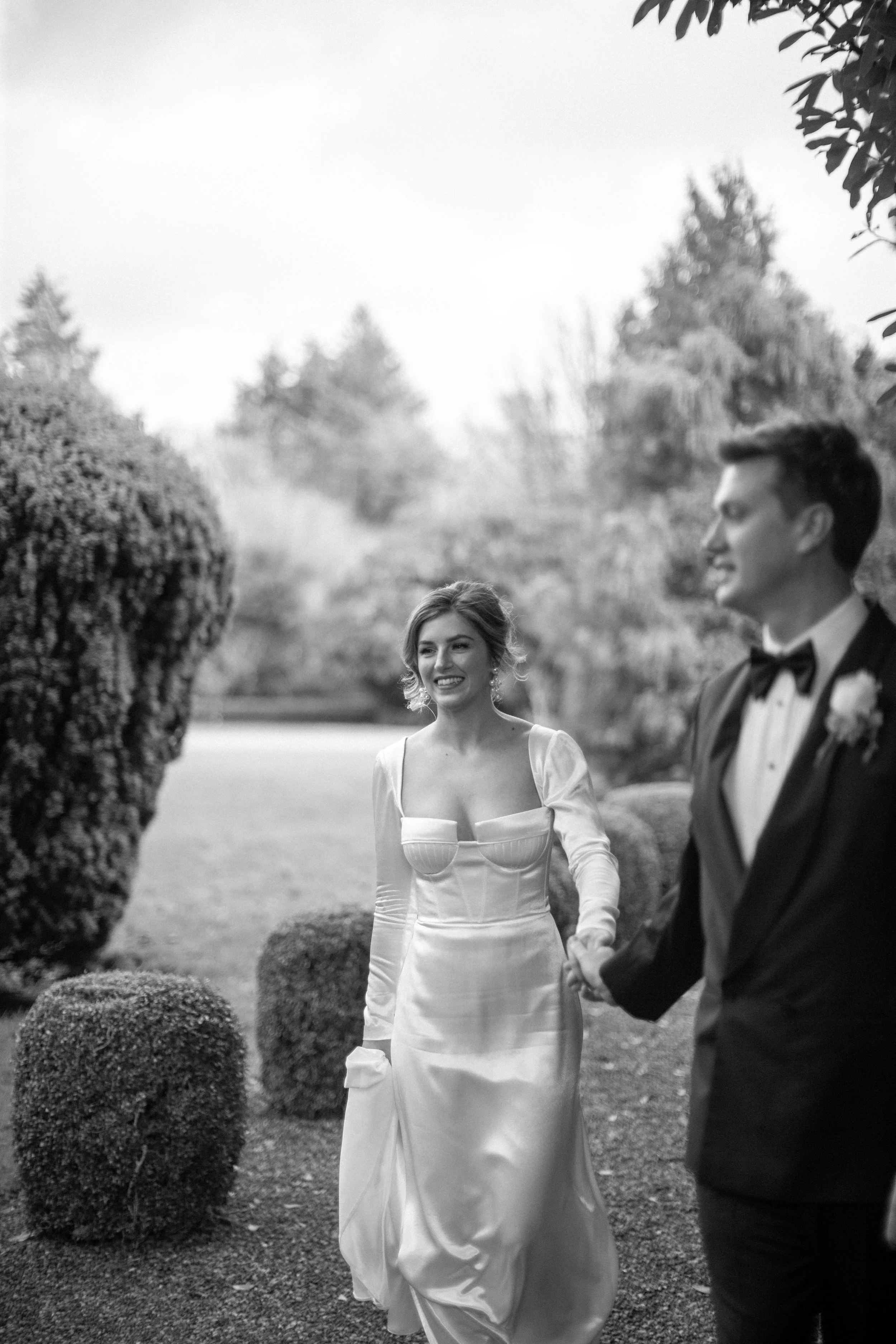 A black and white photo of a woman in a white wedding dress, holding the hand of a man in a tuxedo, outdoors in a garden with bushes and trees in the background.