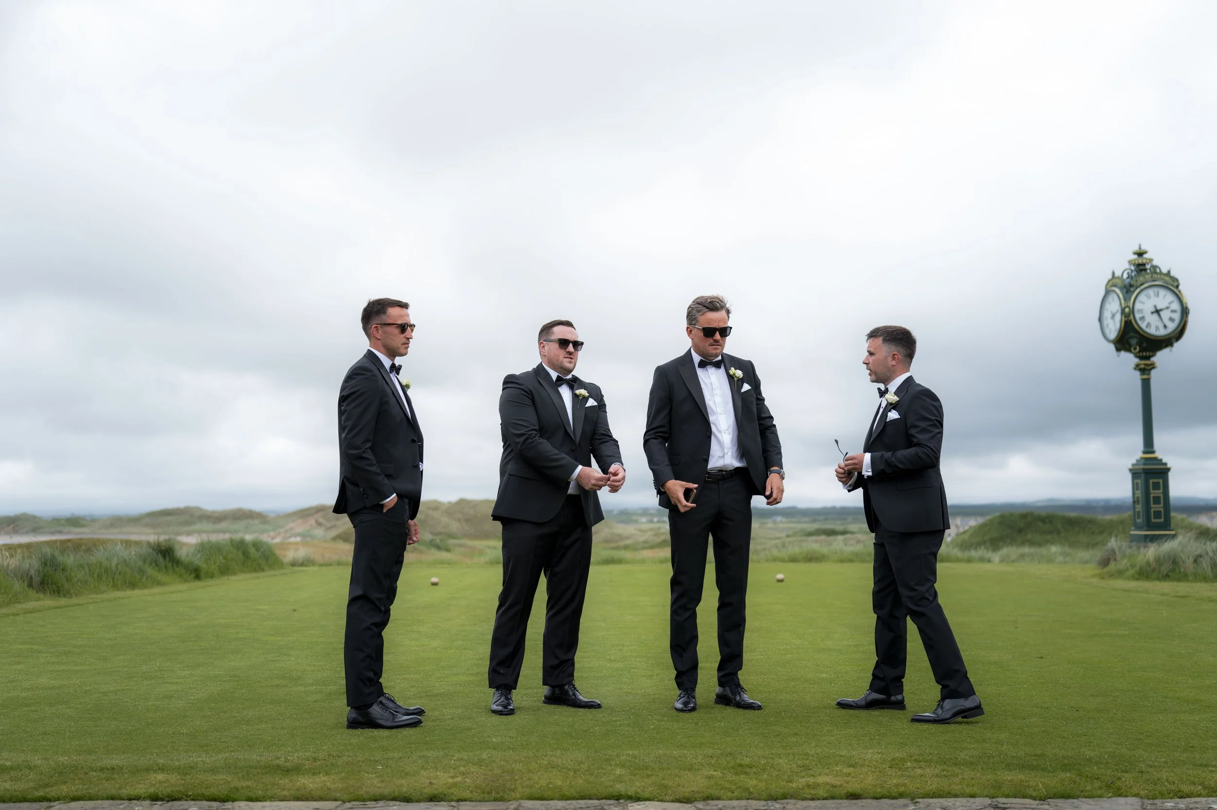 Four men dressed in black tuxedos and sunglasses standing on a golf course with a green landscape and cloudy sky. There is a clock tower in the background.