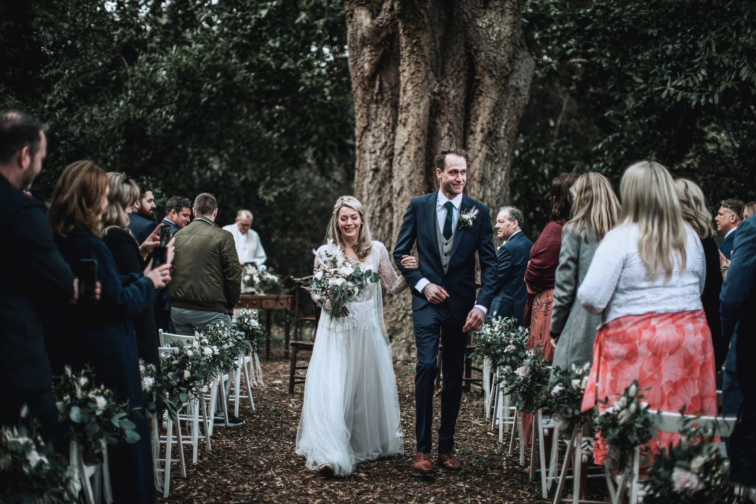 A bride and groom walking down the aisle during their outdoor wedding ceremony in a wooded area with guests watching and taking photos.