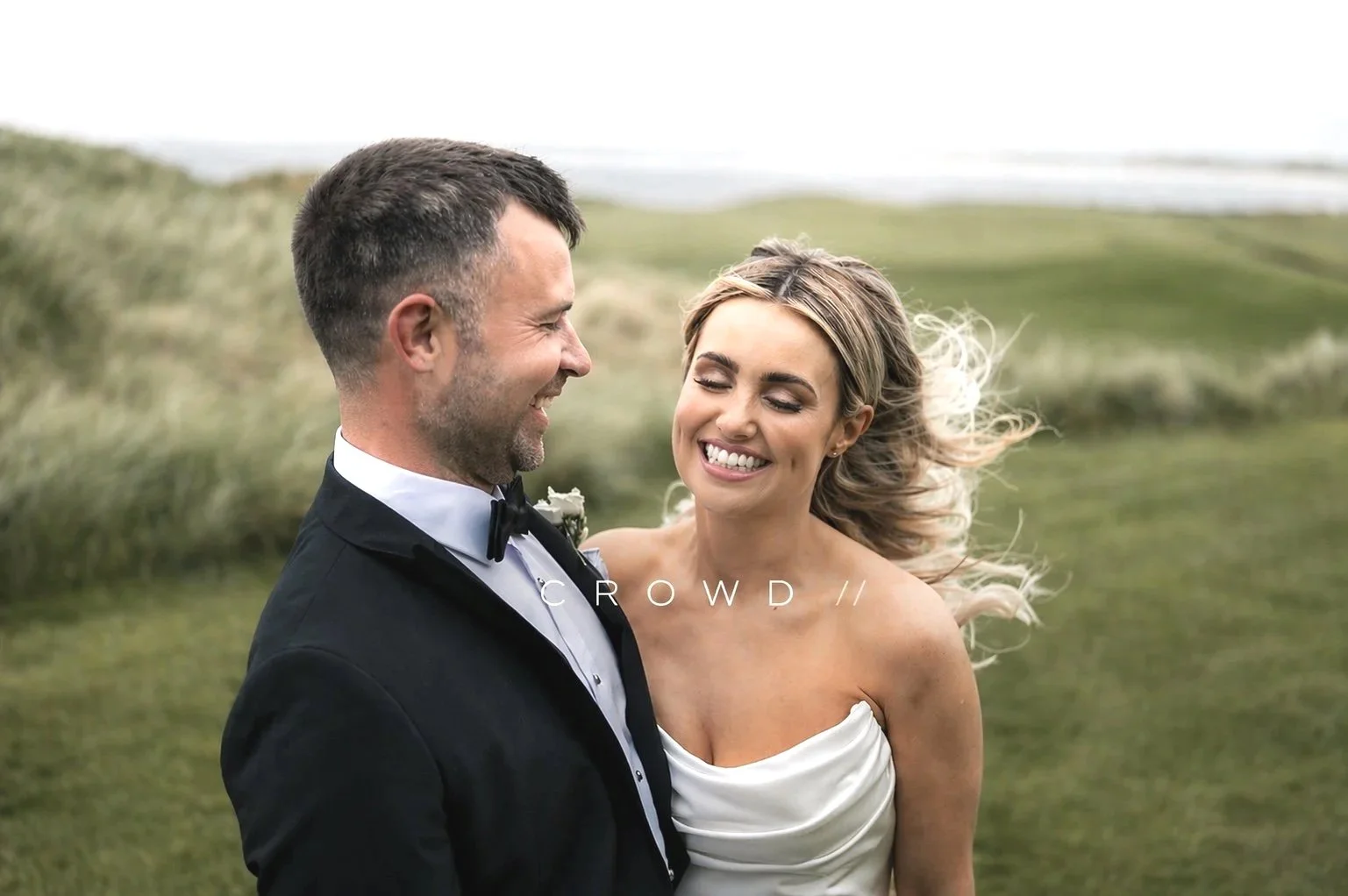 A smiling bride and groom in wedding attire standing outdoors on a grassy area near water, with the groom in a black tuxedo and the bride in a white strapless wedding dress, enjoying a joyful moment together.