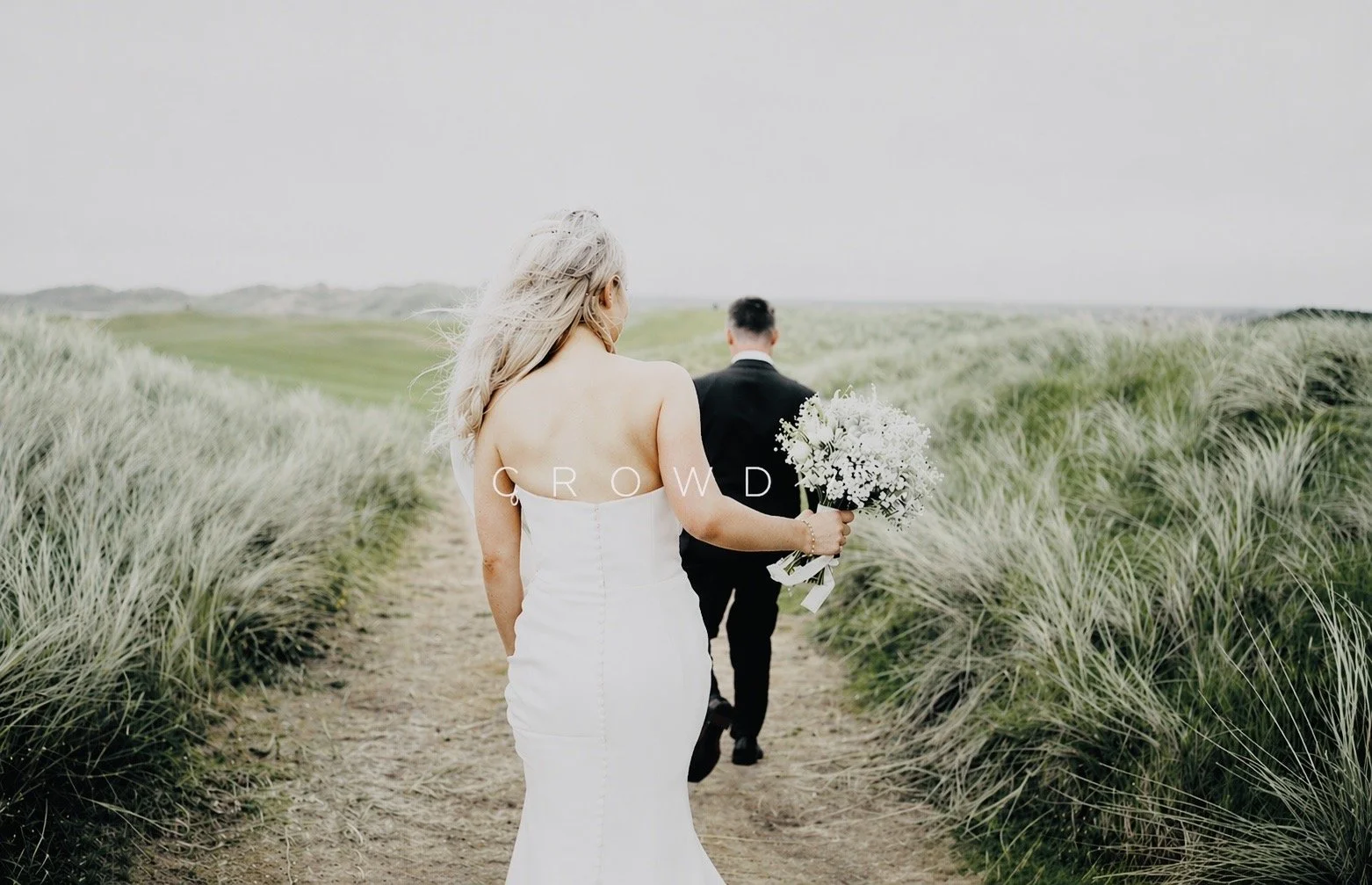A bride in a white wedding dress walking along a dirt path in a grassy field, holding a bouquet of white flowers, with a groom in a black suit walking ahead.