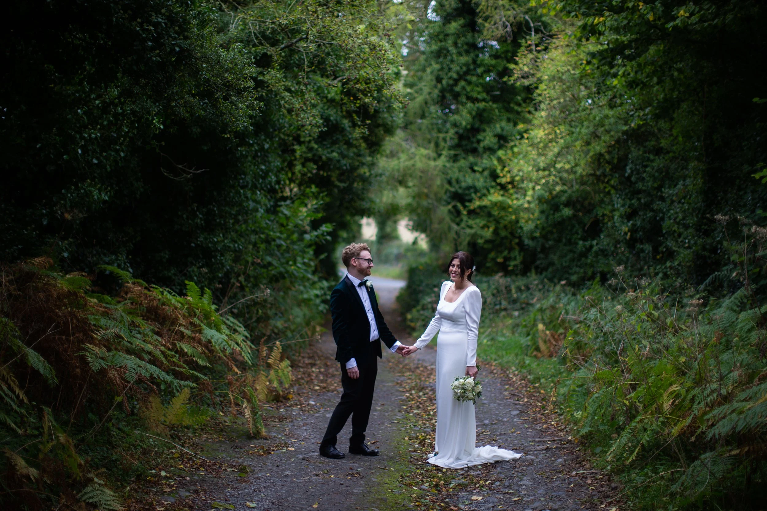A bride and groom are holding hands and standing on a forest trail, smiling at each other. The groom is wearing a black suit and glasses, and the bride is in a white wedding dress holding a bouquet at the Rock of Dunamase Co Laois