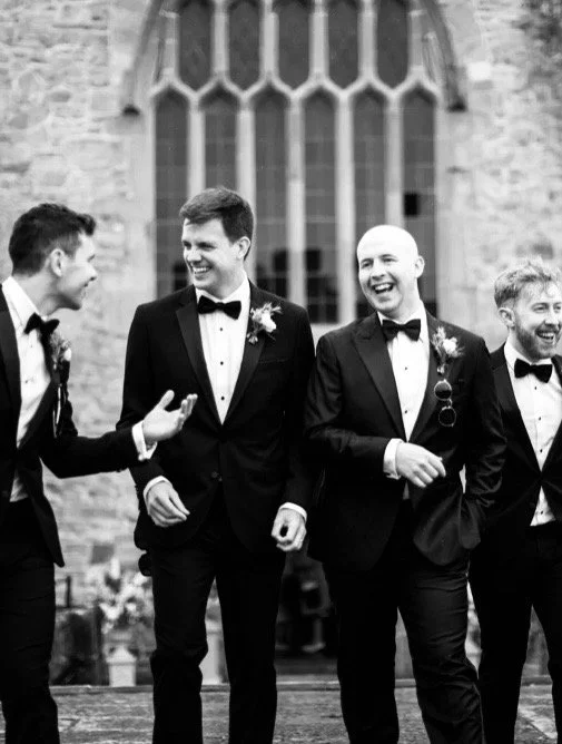 Four men in tuxedos with boutonnières, walking and conversing outside a church, at a formal event.