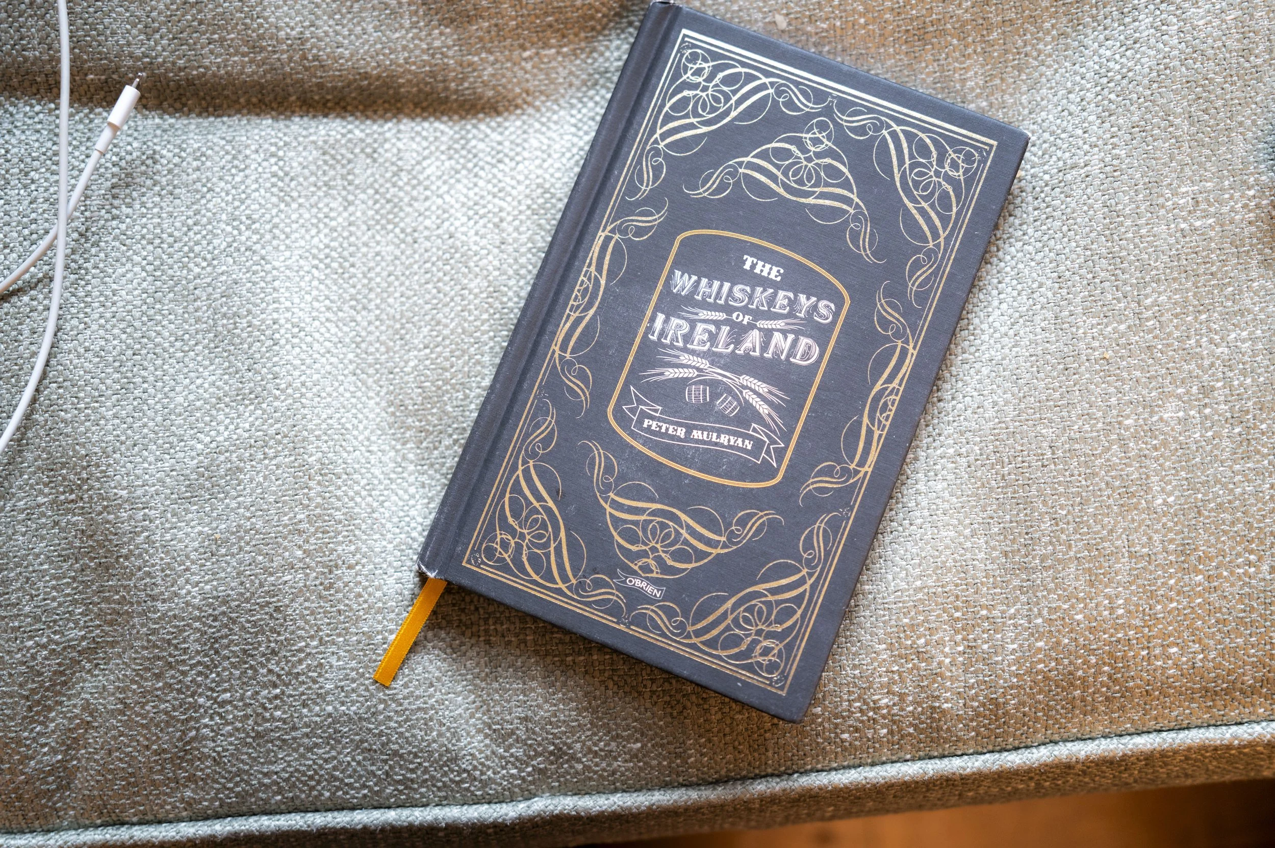 A navy blue hardcover book titled "The Whiskeys of Ireland" by Peter Mulryan, resting on a textured beige fabric surface. The book has gold decorative designs on the cover and an orange ribbon bookmark. A white earphone cable is partially visible to 