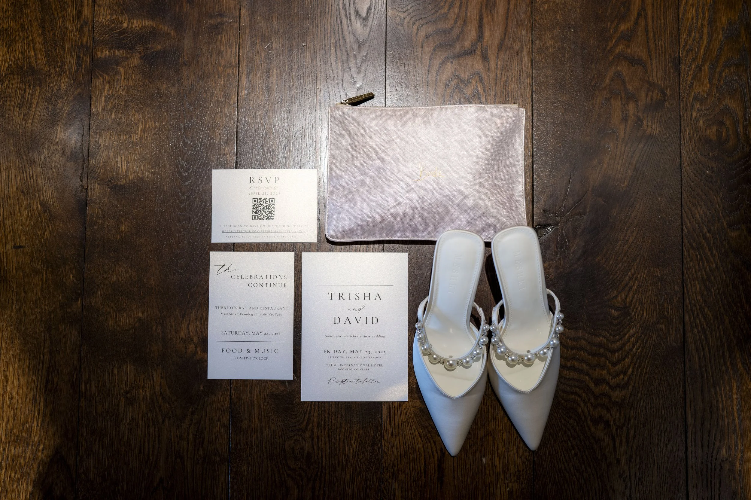 A flat lay of wedding invitation stationery, white pearl decorated shoes, a pink clutch purse, and wedding RSVP cards on a wooden floor.