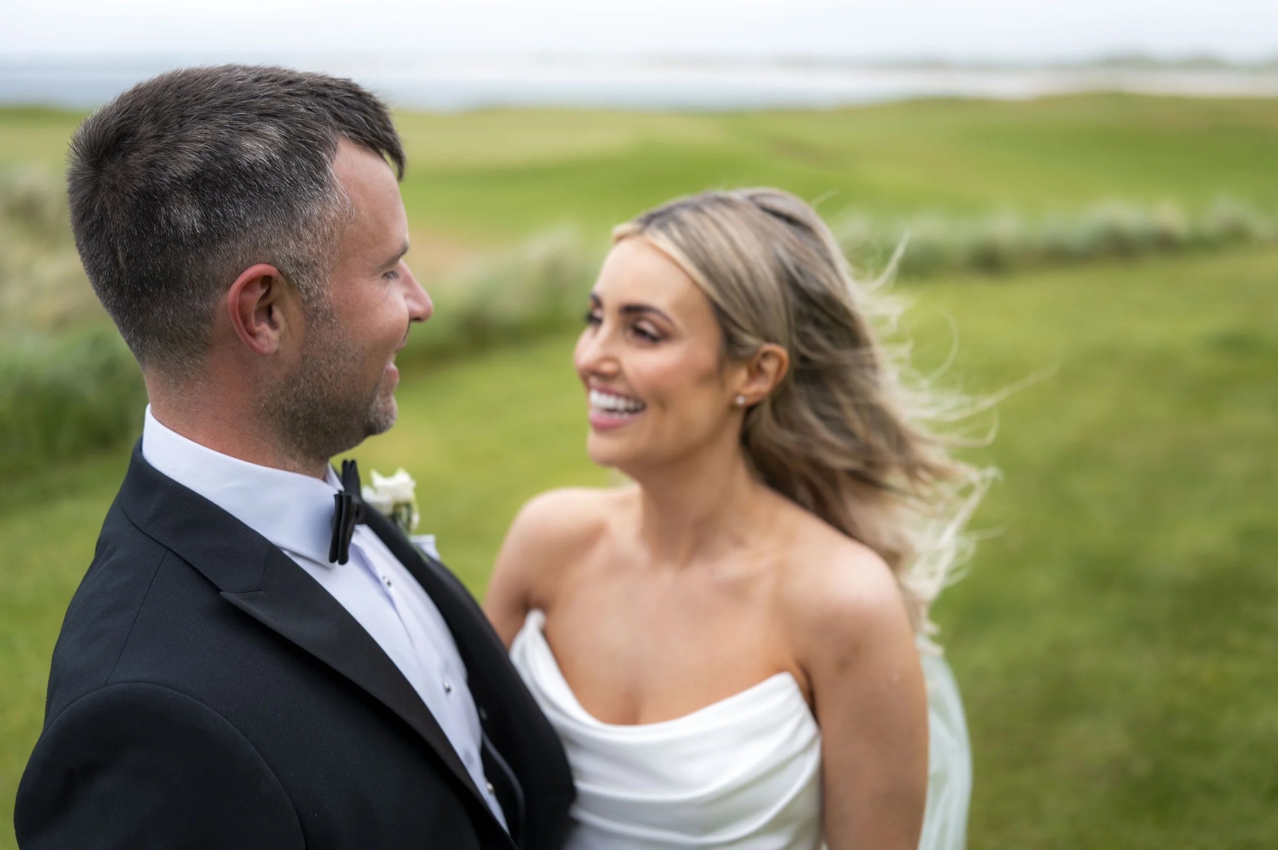 A smiling bride and groom facing each other outdoors with a blurred green landscape background.