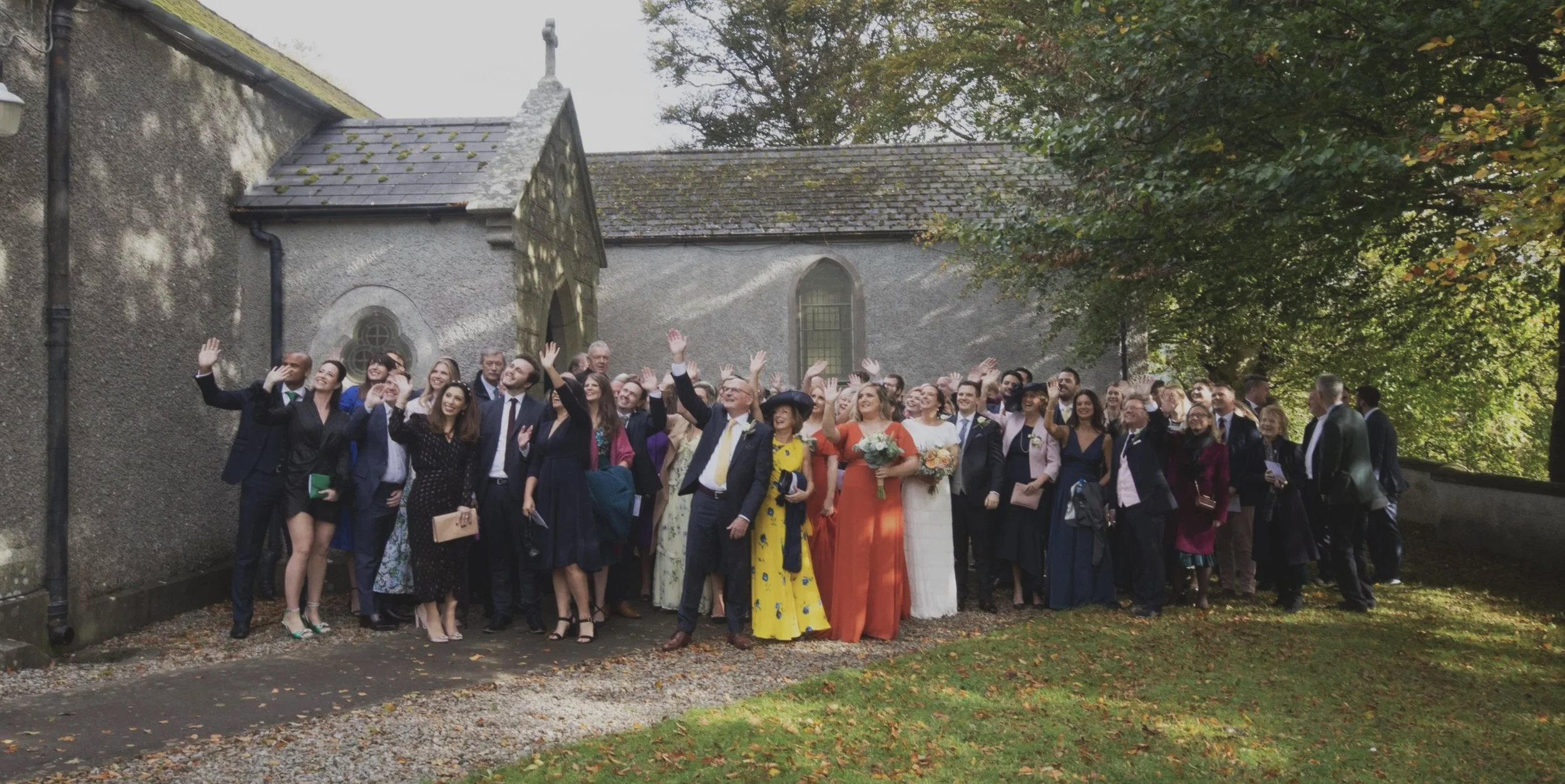 A large group of people, dressed in formal attire, gathered outdoors near an old stone building with trees in the background. The group appears to be celebrating, with many of them smiling, waving, and holding flowers.