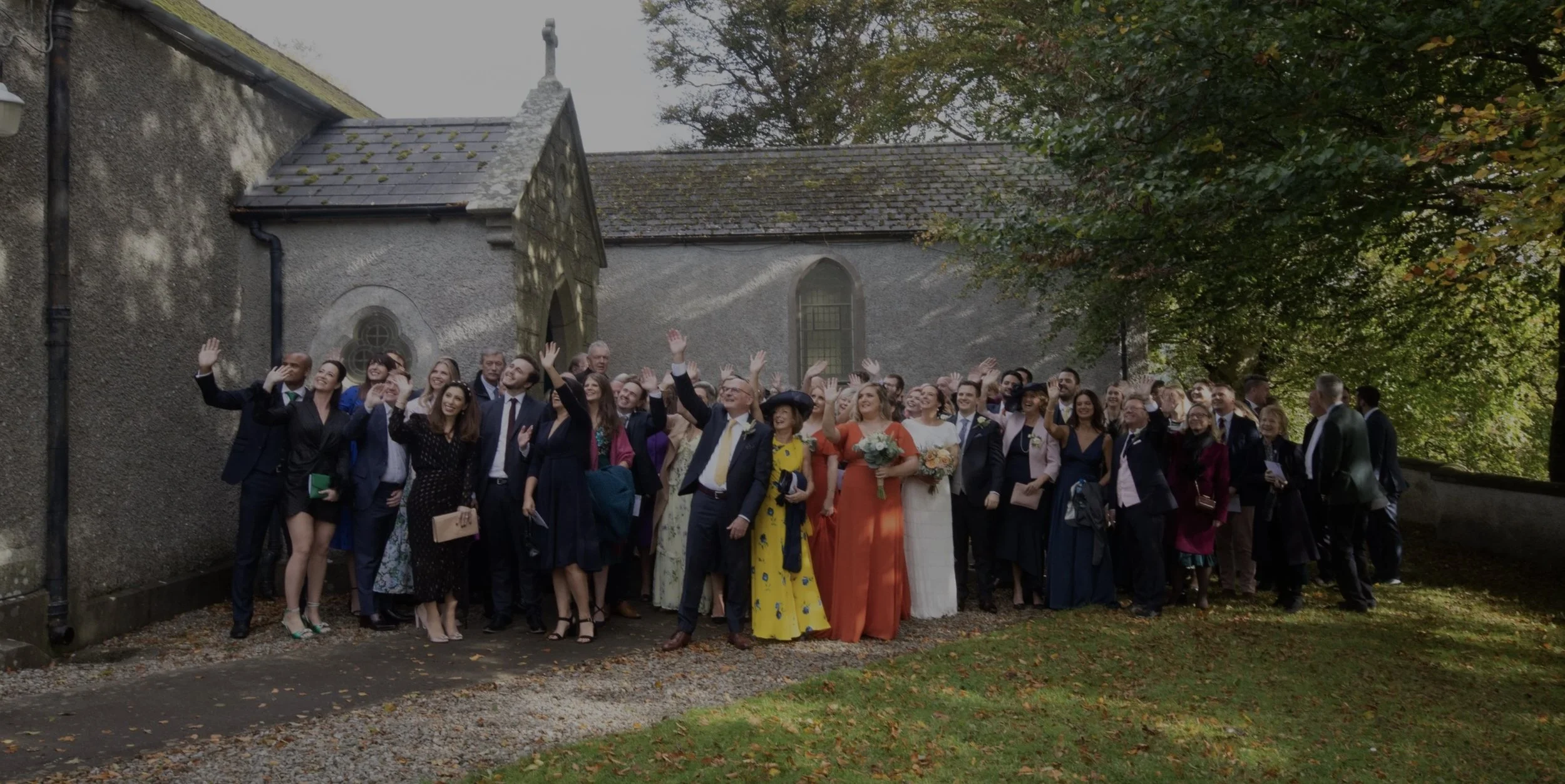 A large group of people gathered outside a church or historic building celebrating a wedding, with many guests waving and smiling, some holding flowers, on a partly cloudy day with trees in the background.