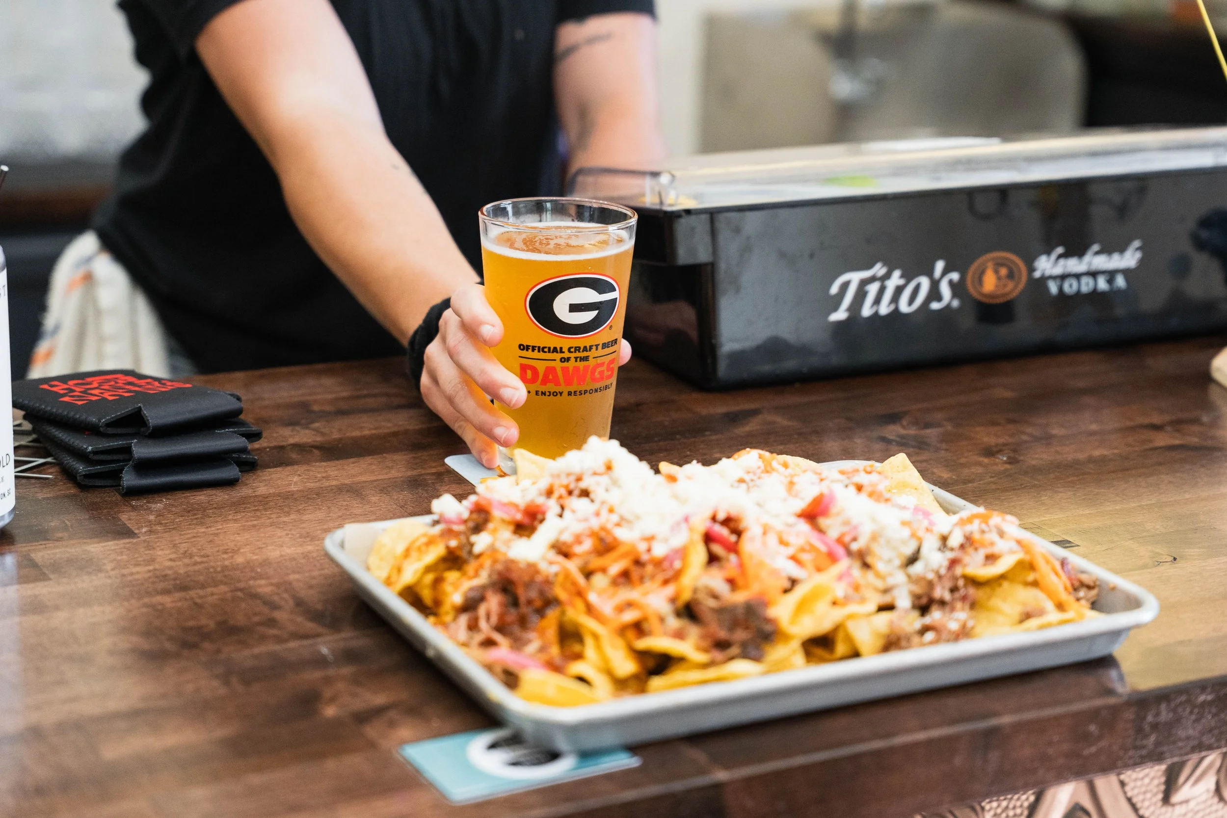 Hootenanny's bar a glass of craft beer with a 'G' logo and the words 'Official Craft Beer of the DAWGS' on it, standing at a bar with a tray of nachos topped with cheese, meats, and vegetables, and a black Tito's Vodka box in the background.