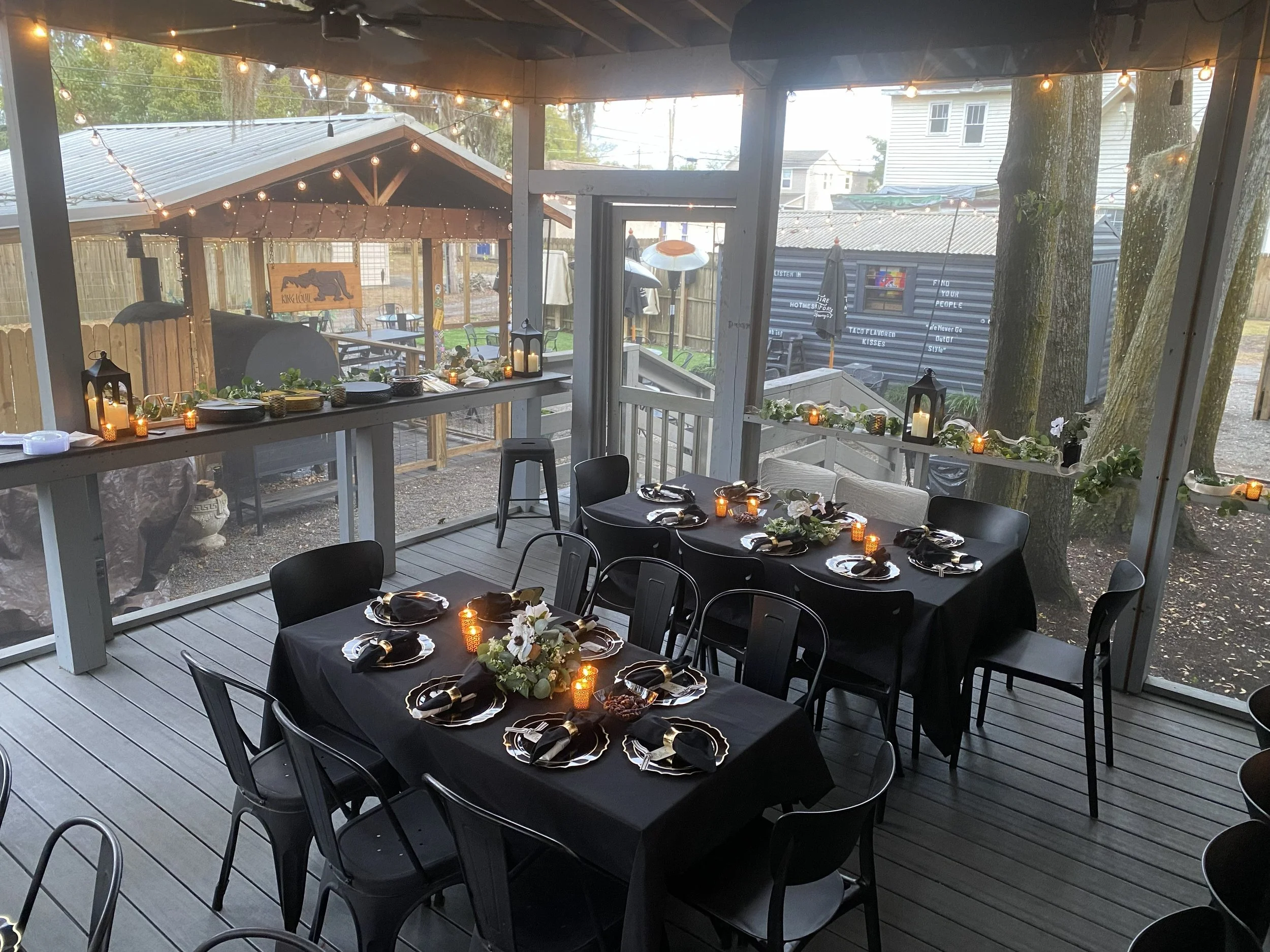 Outdoor covered dining area at Hootenanny's decorated for an event with black tablecloths, floral centerpieces, black napkins, candles, and string lights, with an outdoor view of trees, neighboring houses, and a fence.