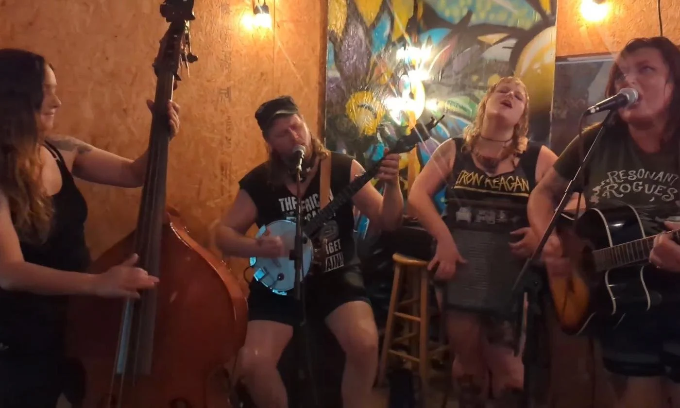 Four women performing music on a small stage in a cozy venue, playing various instruments including an upright bass, a banjo, and an acoustic guitar, with colorful artwork and warm lighting in the background.