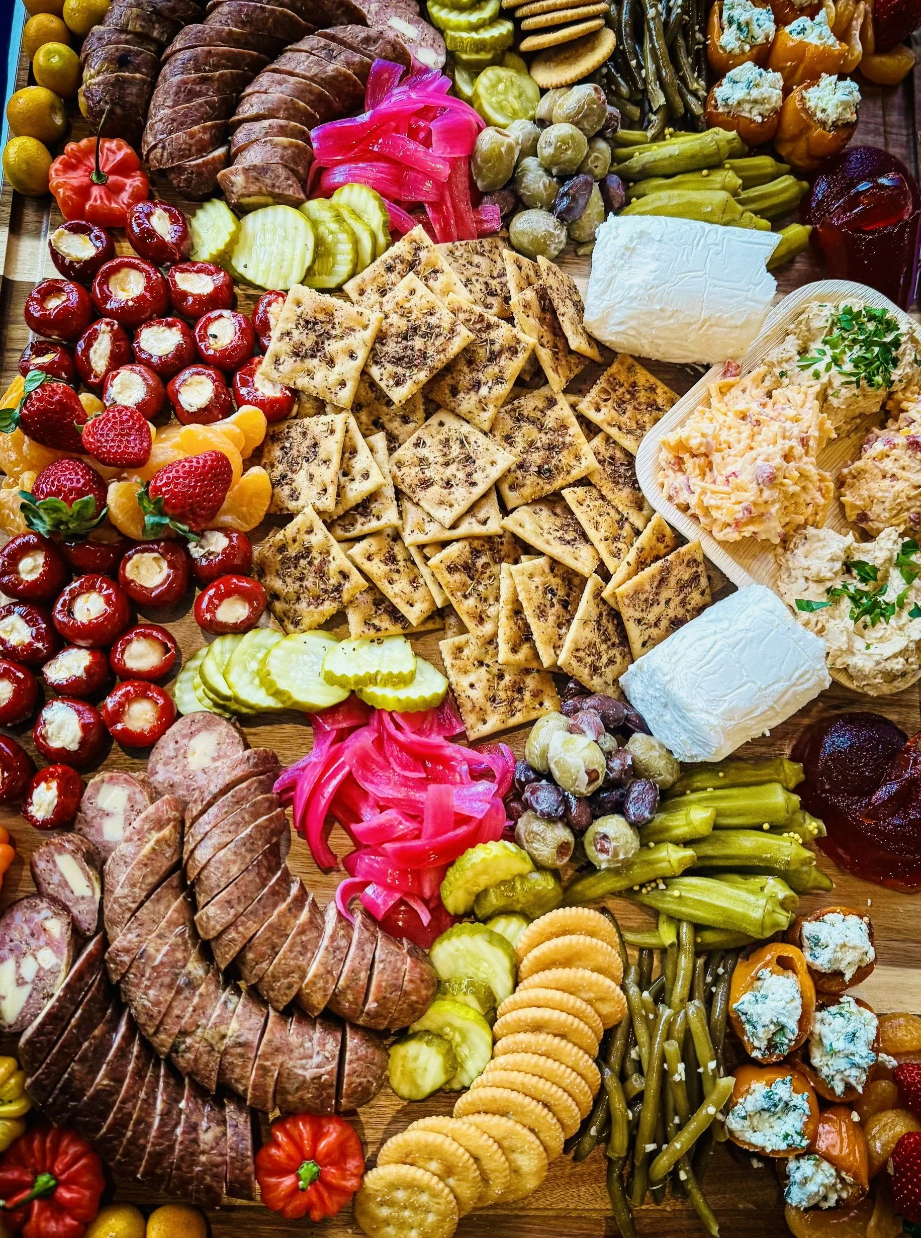 A charcuterie board featuring sliced roasted meats, mixed pickles, sliced vegetables, crackers, cheeses, nuts, and various garnishes arranged on a wooden surface.