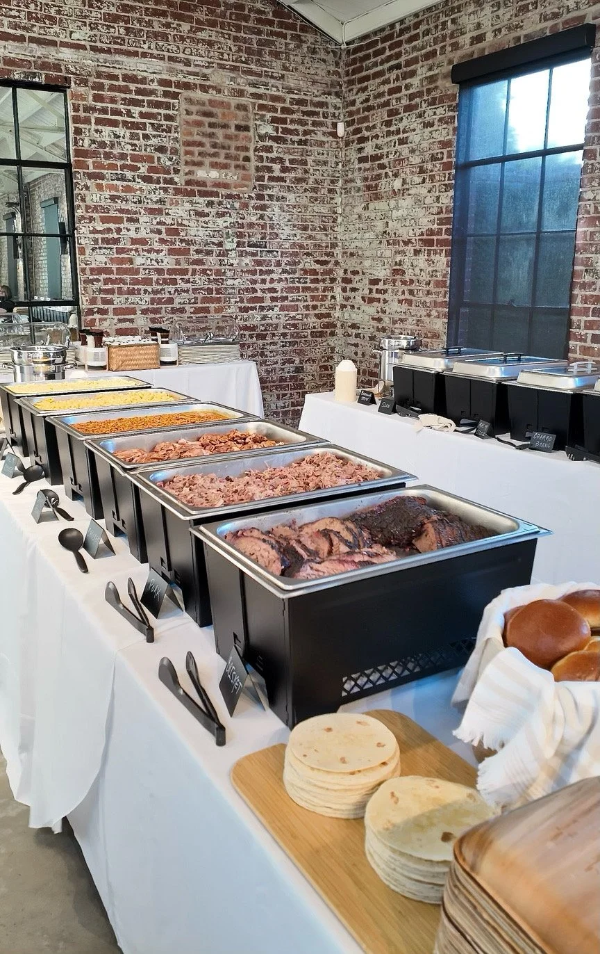 Buffet table with trays of cooked meats, tortillas, and buns, in front of a brick wall and window, with serving utensils and condiments prepared by the Hootenanny's BBQ catering team.