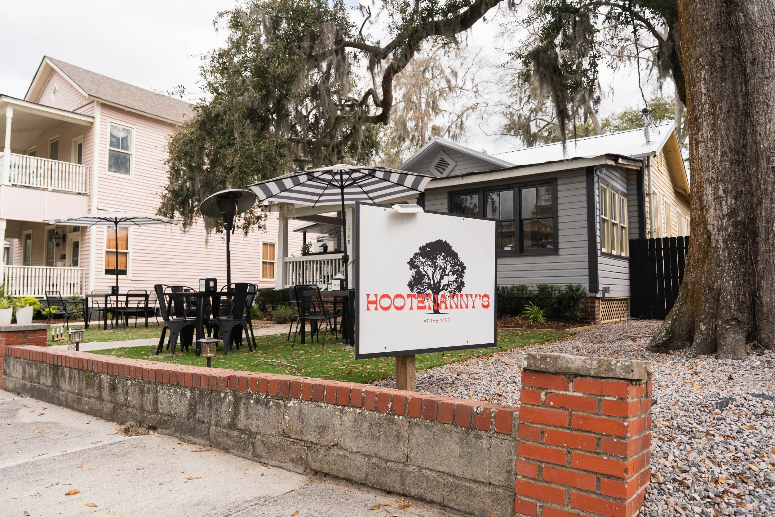 Outdoor seating area in front of a building with a sign for Hootenanny's at the yard, with black tables and chairs, umbrellas, a brick and concrete wall border, and large trees in the background.