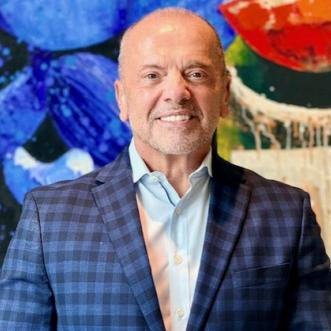 Man with a light blue shirt and checkered blazer smiling in front of a colorful abstract background.