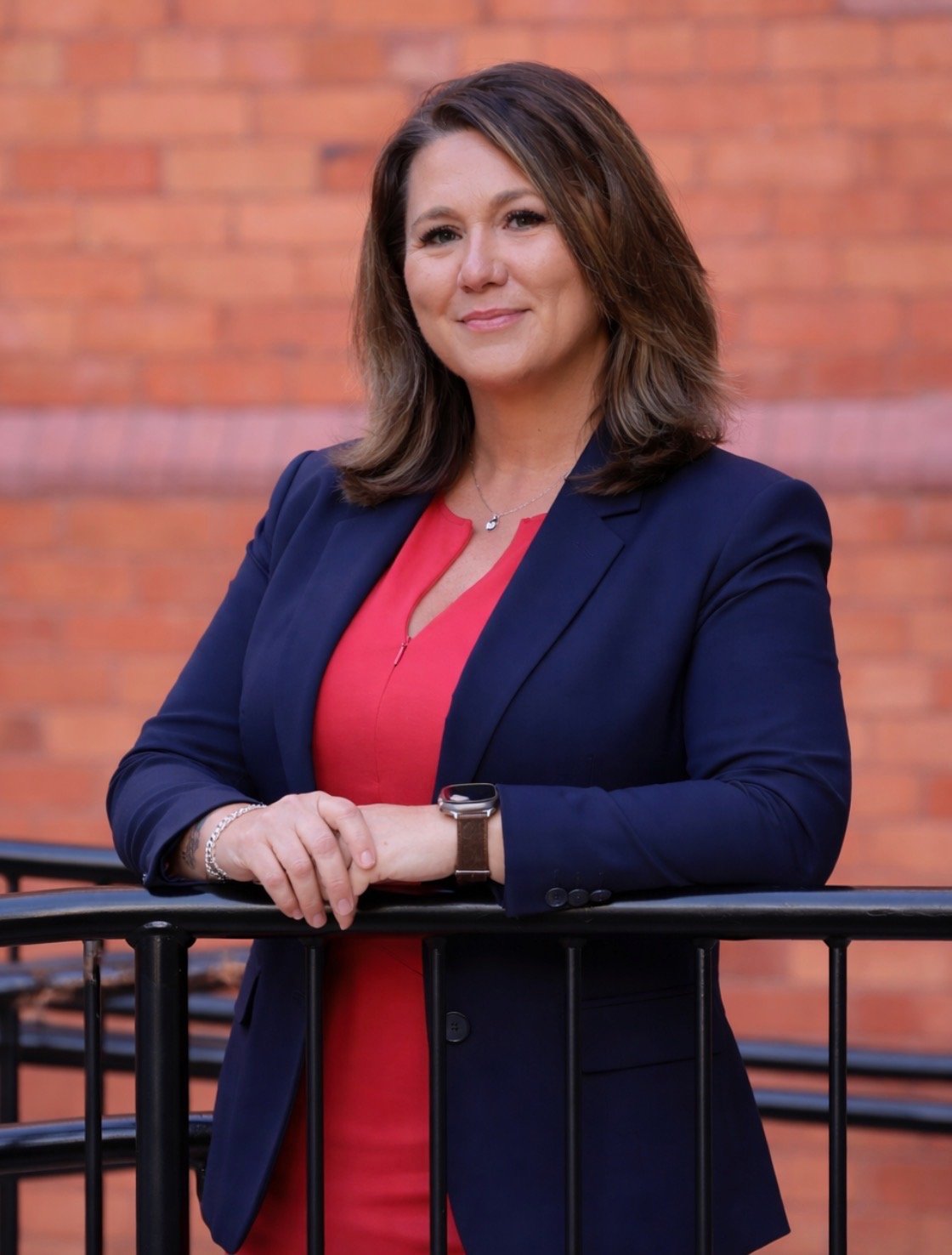 A woman with shoulder-length brown hair wearing a navy blazer over a red top, standing outdoors in front of a brick wall, leaning on a black railing with her arms crossed and smiling.