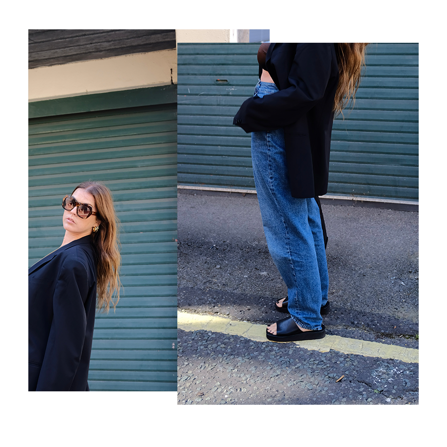 A woman with long brown hair wearing sunglasses, a black blazer, and earrings posing outdoors in front of a green corrugated metal garage door.