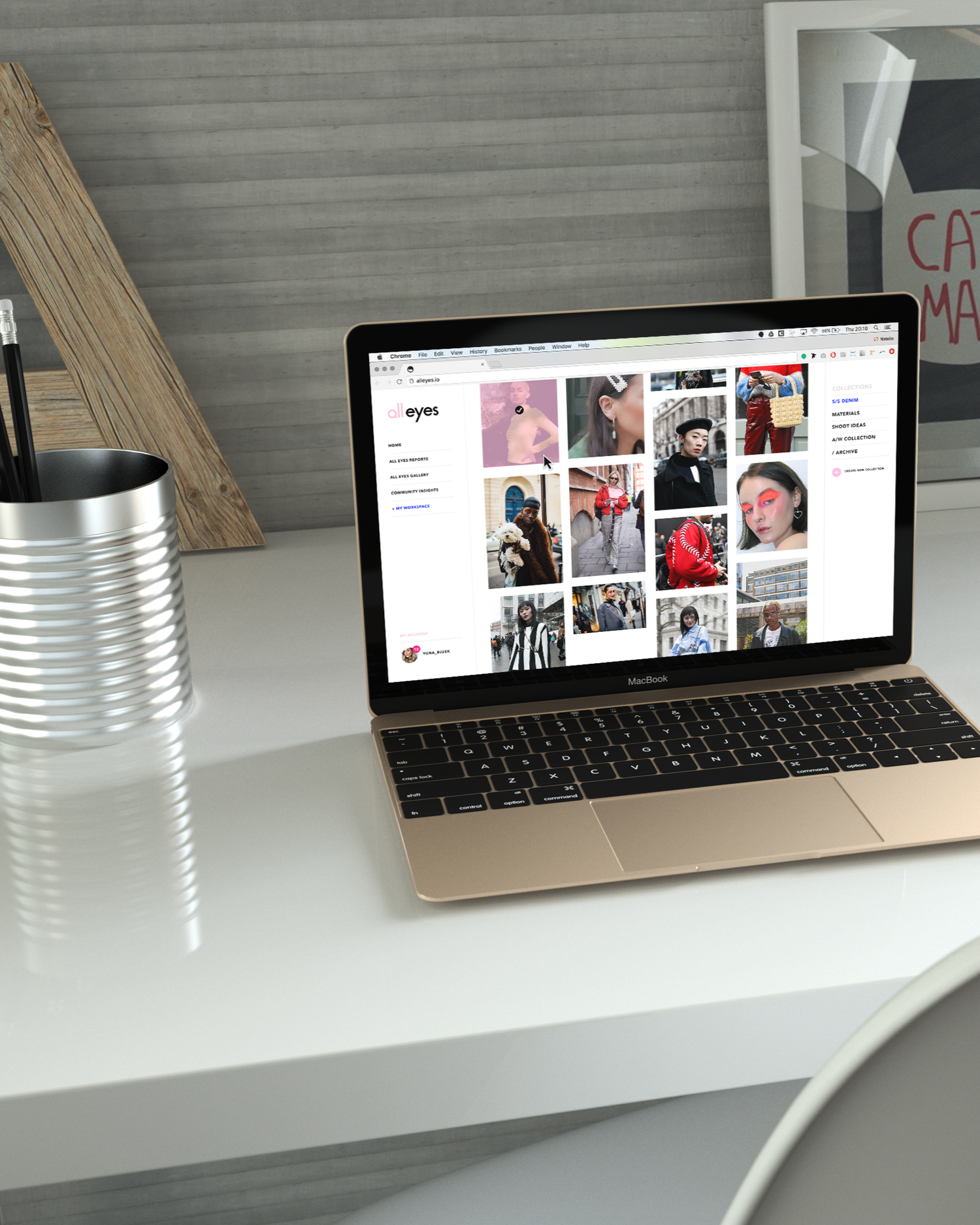 A silver MacBook laptop displaying a website called 'all eyes' with various fashion photos, placed on a white desk with a metal pencil holder and a wooden plank behind it.