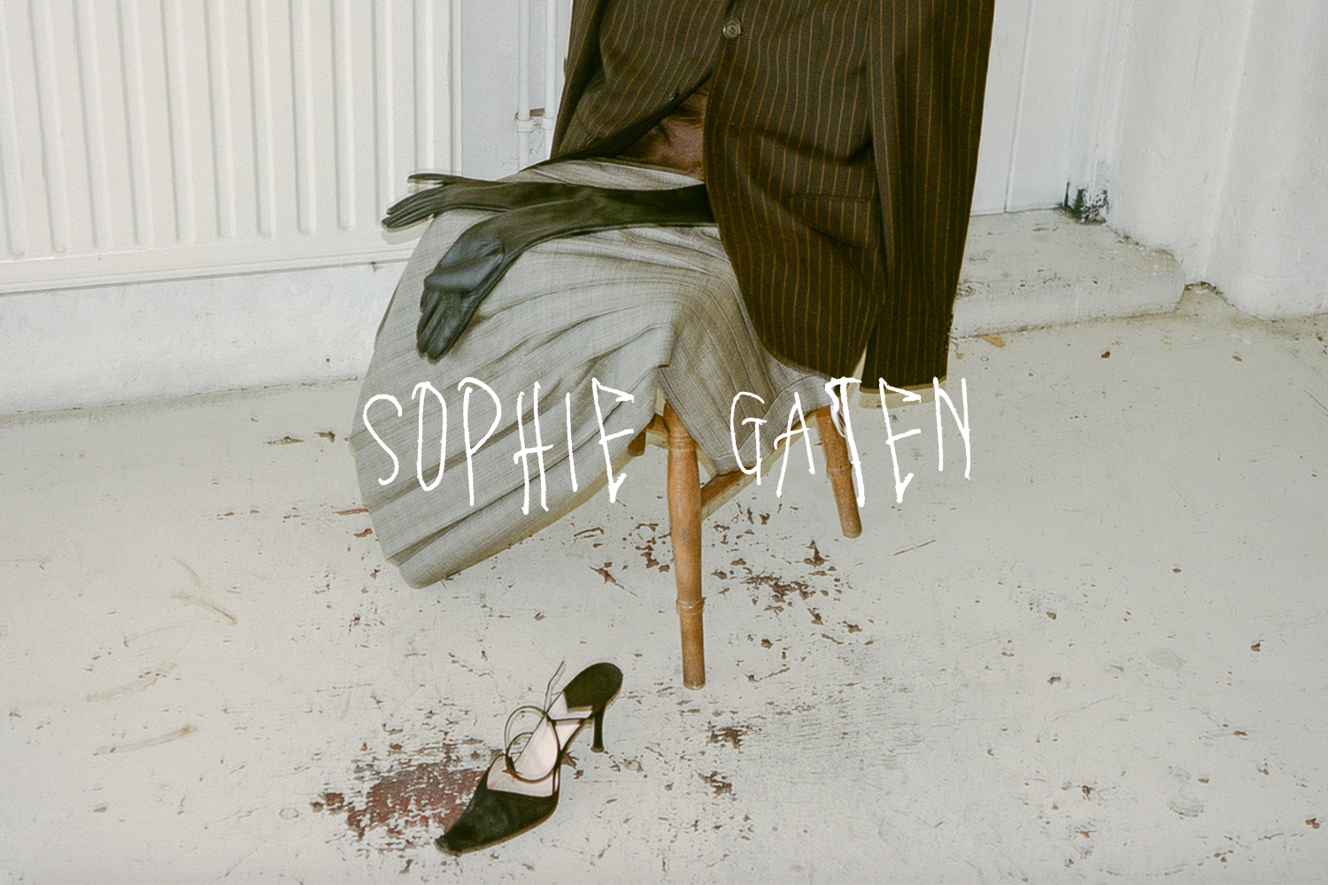 An image of a striped suit jacket plaid pants on a wooden chair. A black high-heeled shoe is on the floor in front of the chair. The background shows a white wall and a gray painted floor with paint chips and stains. The text 'SOPHIE GATEN' is written across the image.